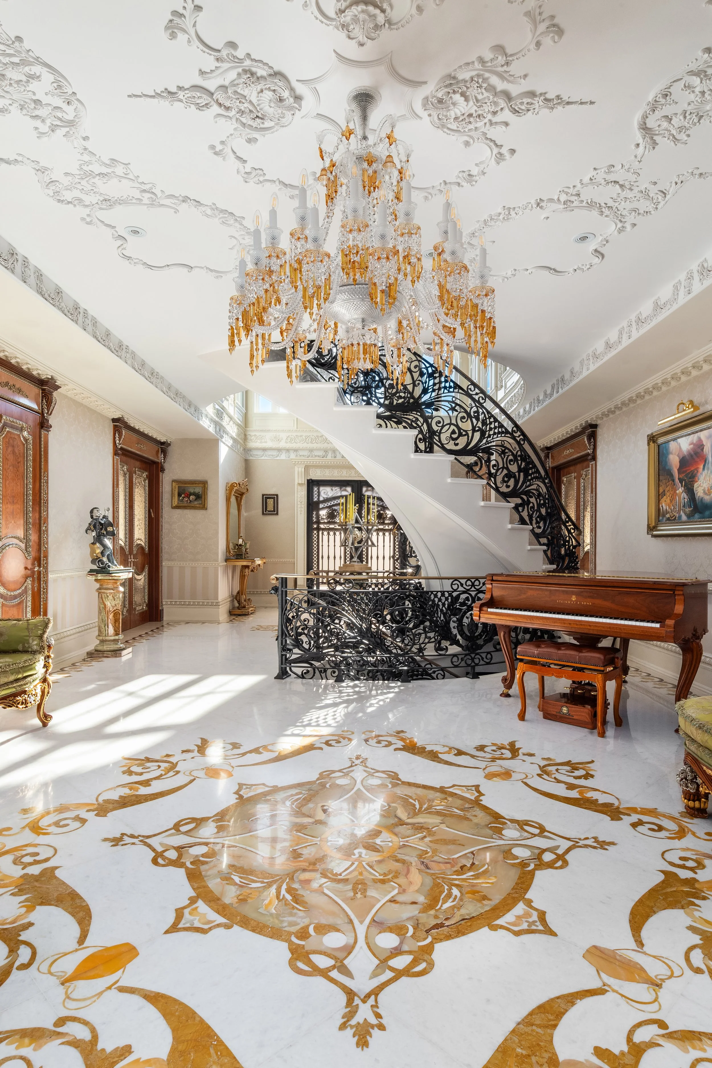 Opulent interior space featuring a large crystal chandelier, ornate ceiling medallions, a curved staircase with wrought iron railing, a grand piano, and decorative furniture and artwork.