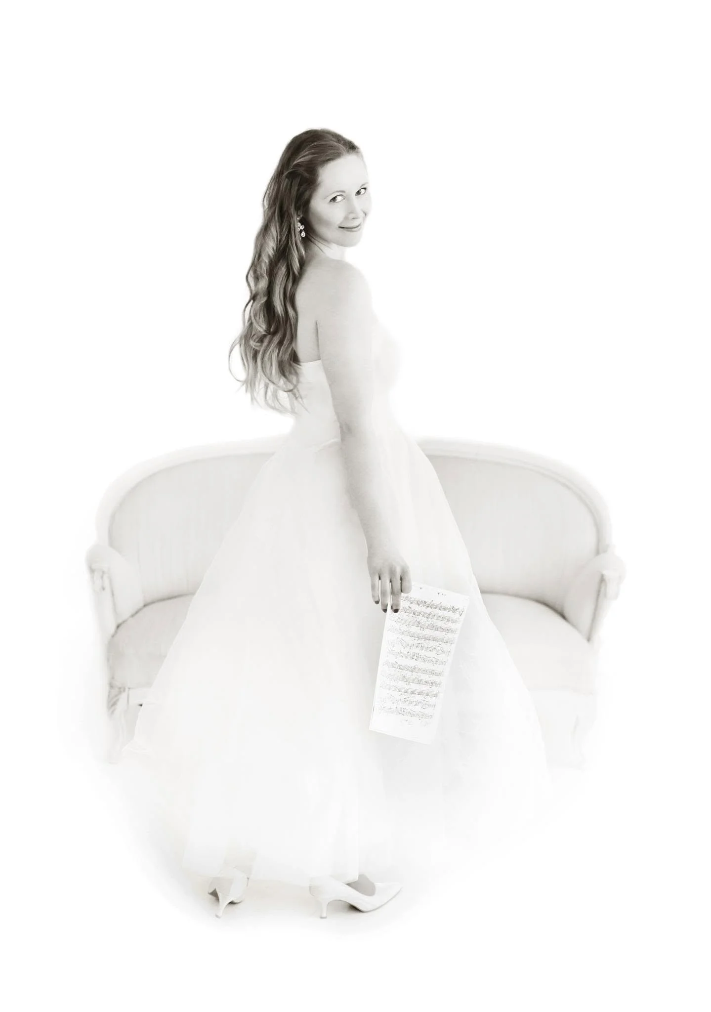 A woman with long wavy hair wearing a strapless dress and earrings, standing next to a curved white sofa, holding a piece of paper.