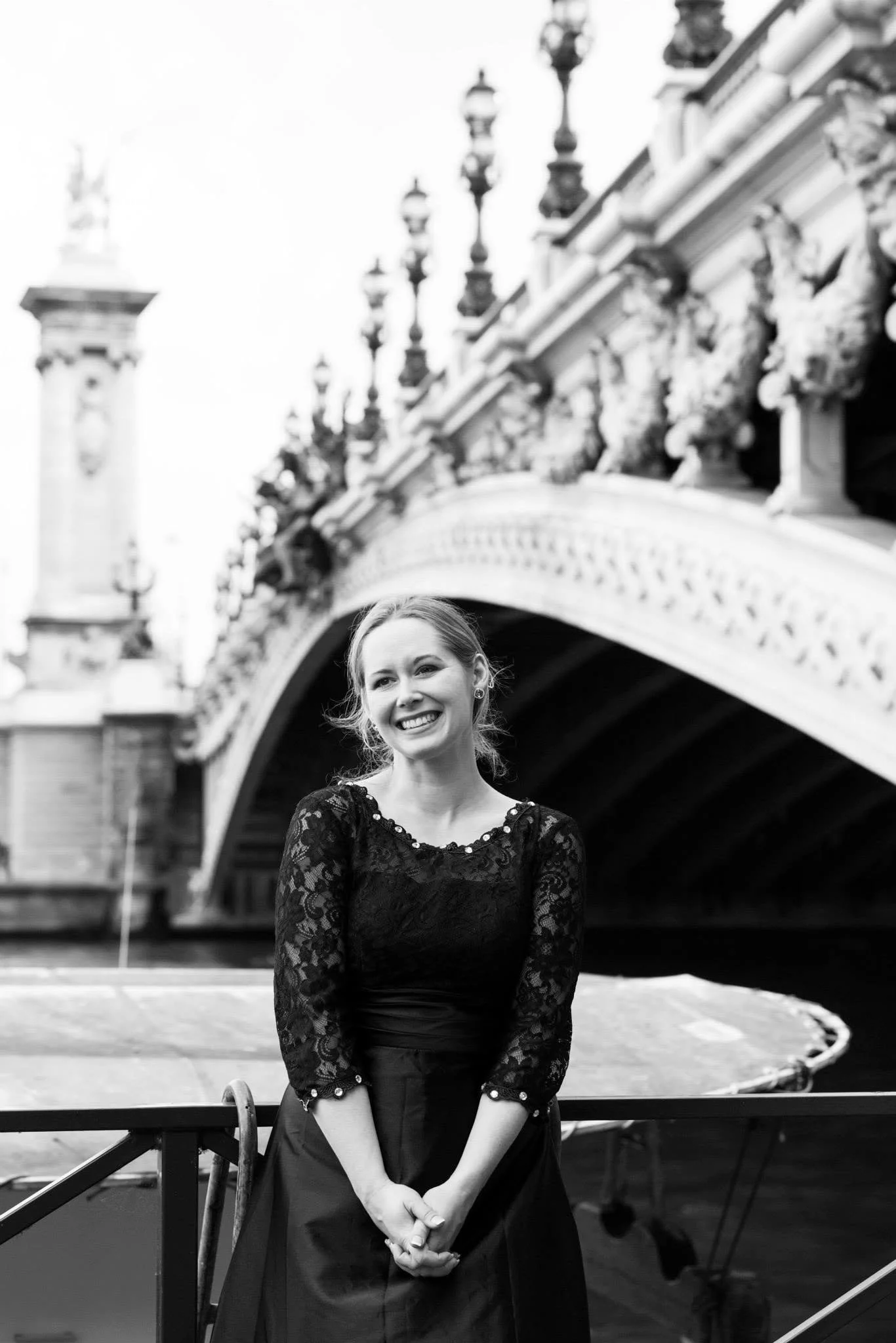 Black and white photo of a woman standing in front of a bridge over a body of water, smiling, wearing a black lace long sleeve top and a black skirt, with a cityscape background.