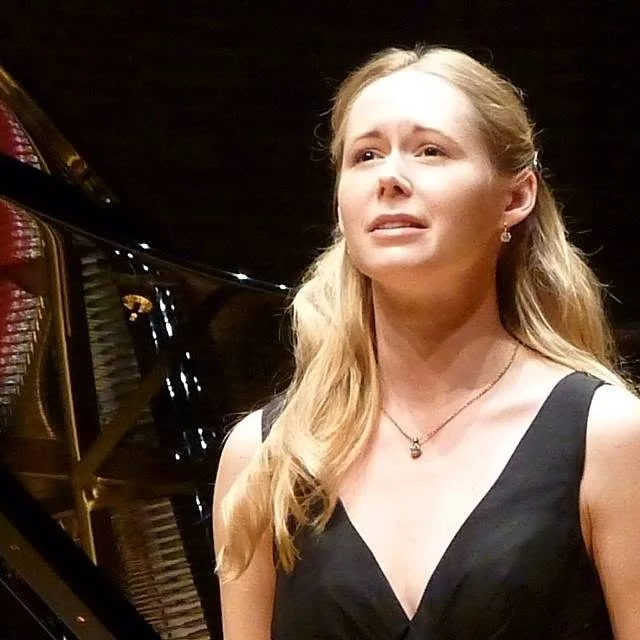 A young woman with long blonde hair wearing a black dress and necklace, appearing emotional or distressed, standing near a grand piano.