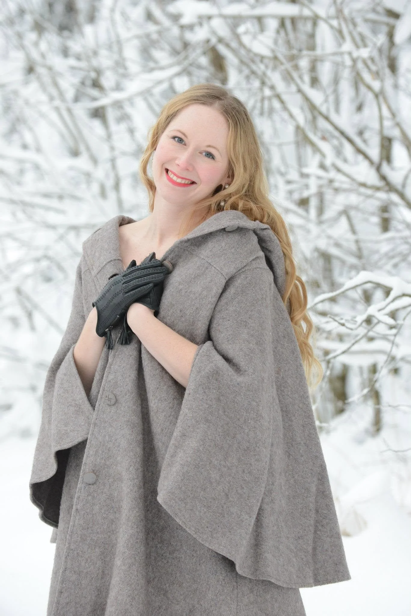 A smiling woman with long wavy ginger hair, wearing a gray coat and black gloves, standing outdoors in a snowy forest.