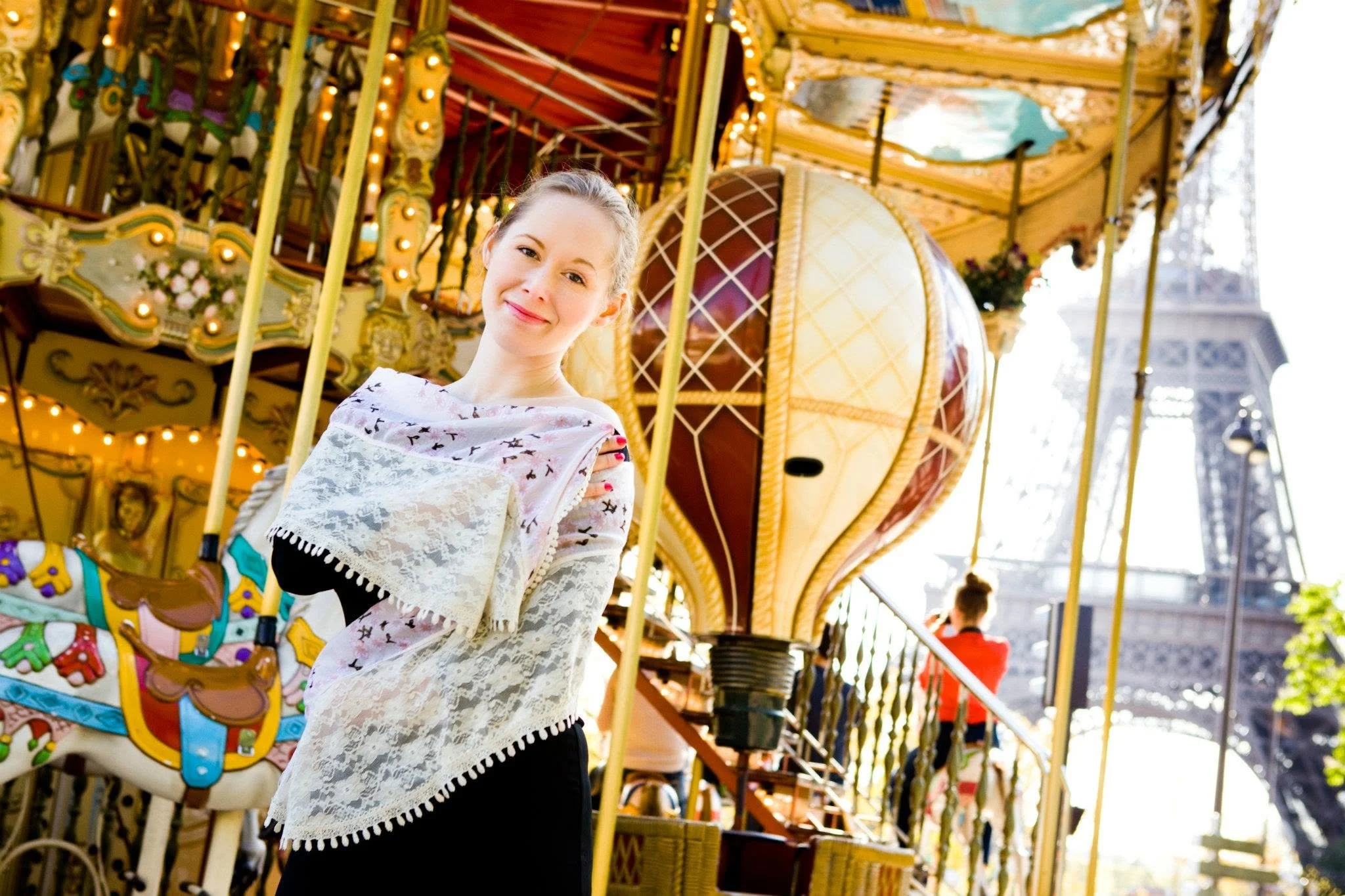 A young woman with a gentle smile standing in front of a colorful carousel at a fairground, with bright lights and spinning rides in the background.