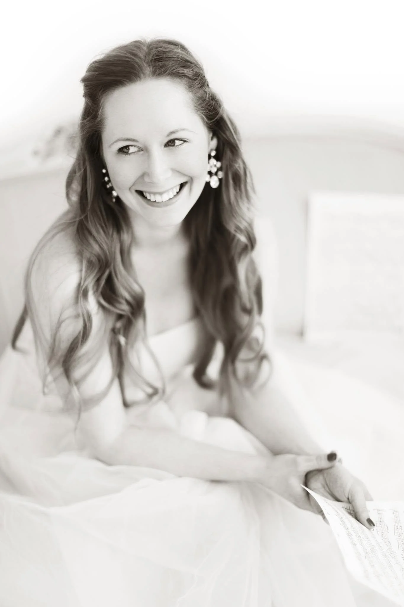 Black and white photo of a woman with long, wavy hair smiling and wearing earrings, sitting on a bed with white bedding.