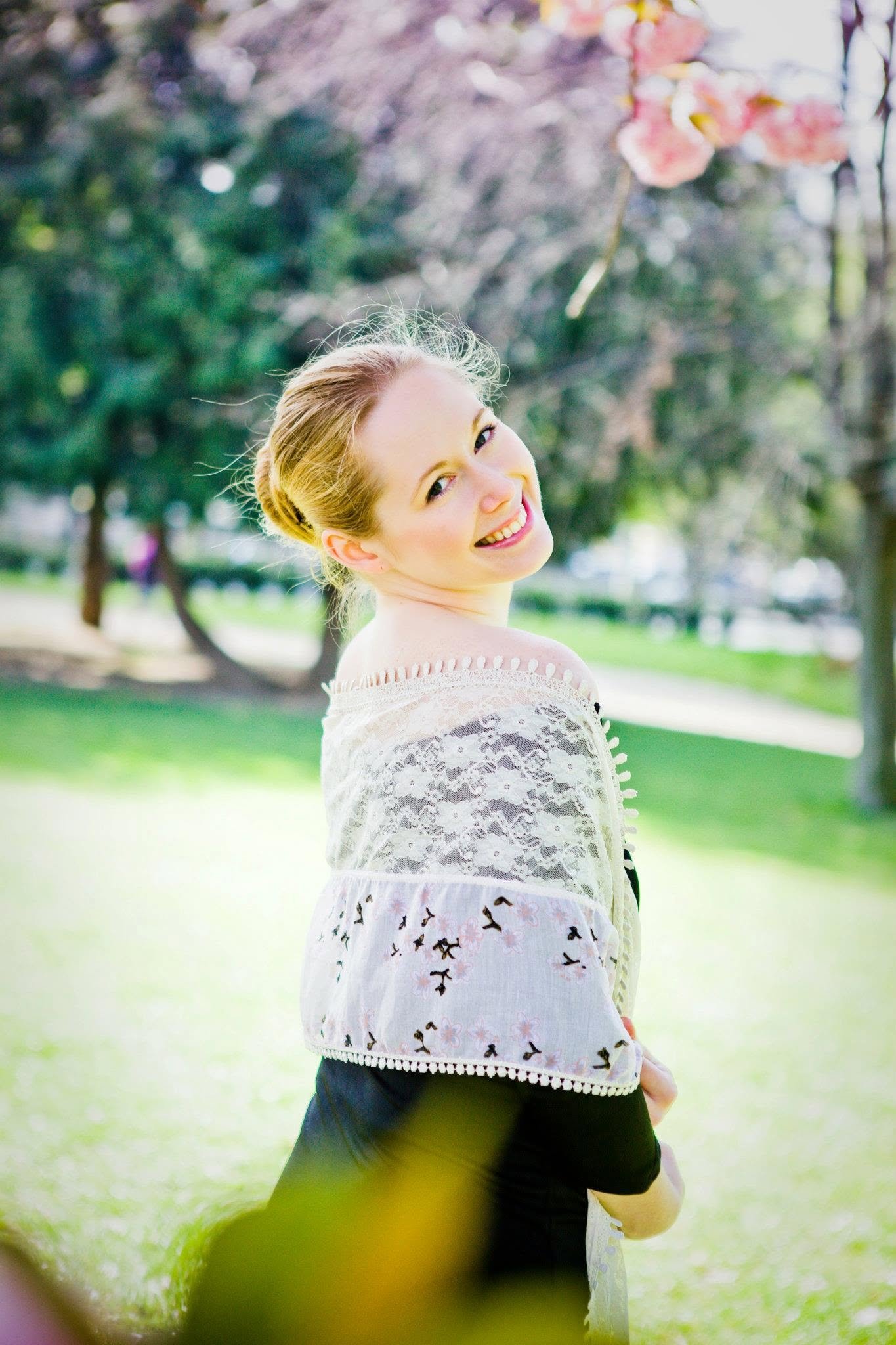 A smiling woman with blonde hair in an updo, wearing a white lace and embroidered top, standing outdoors in a park with trees and soft sunlight.