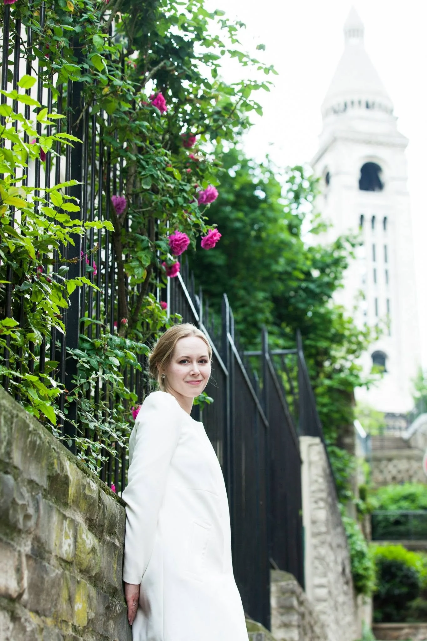 A woman in a white dress standing by a stone wall with a black metal fence that has pink flowers, with a church steeple in the background.