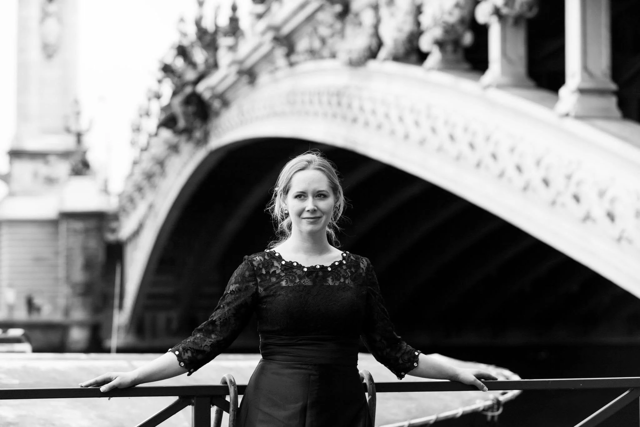 A black and white photo of a woman with light-colored hair standing outdoors under a bridge, wearing a lace dress with three-quarter sleeves, her arms resting on a railing, looking slightly to the side.