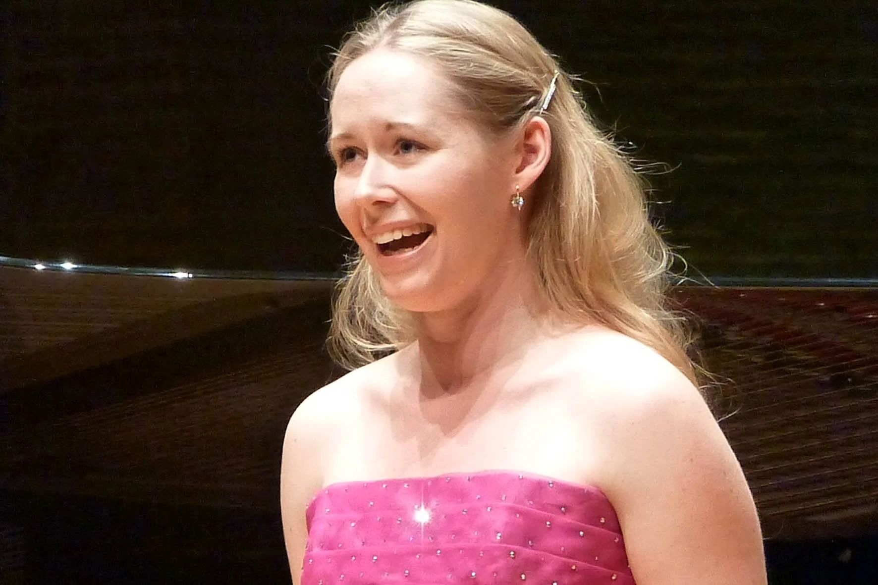 A woman wearing a pink strapless dress with rhinestones, smiling and looking to her left, with earrings and a hair clip, against a dark background.