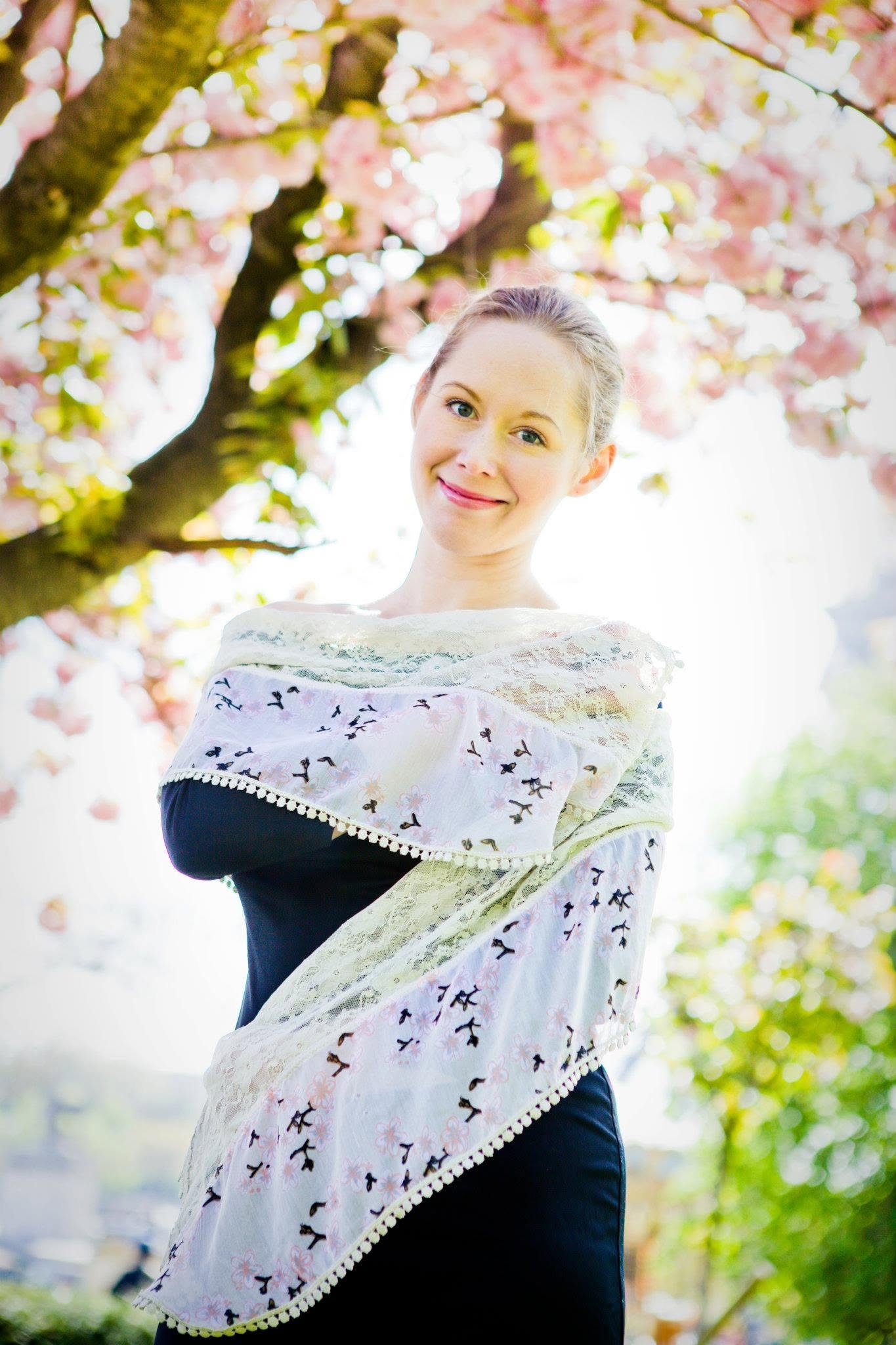 A smiling woman standing outdoors under a blooming pink cherry blossom tree, wearing a white shawl with black and pink embroidery over a black dress.