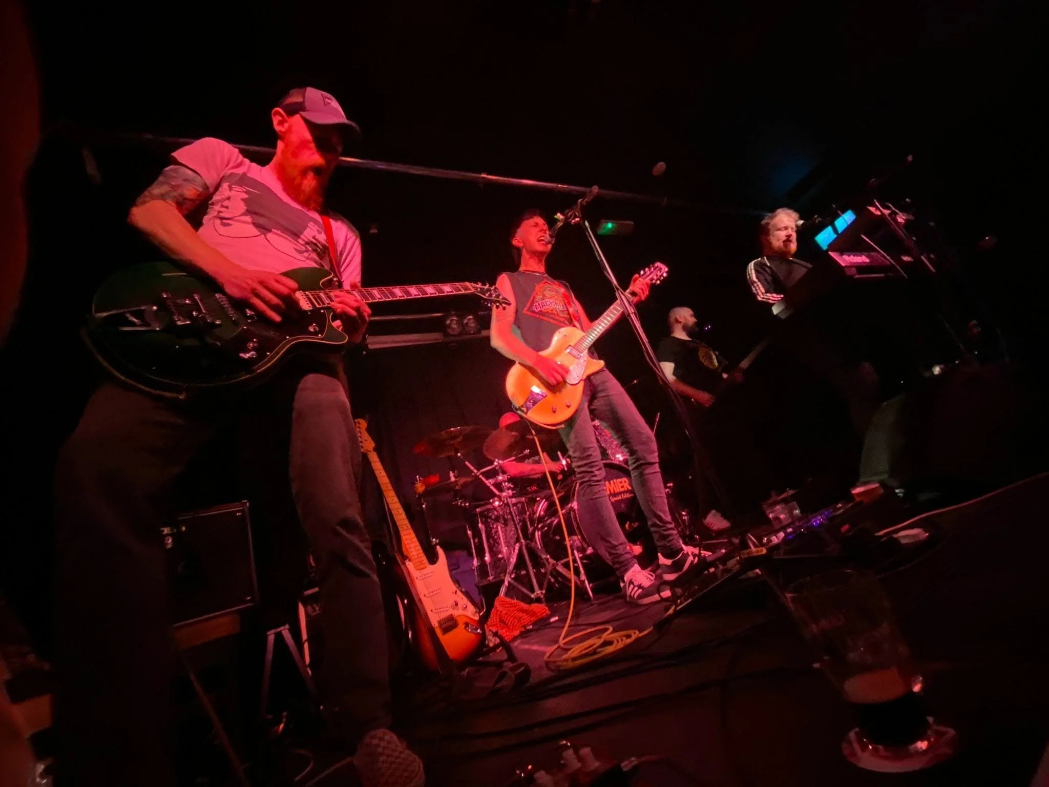 A band performing on stage with three visible musicians playing guitars and drums under red lighting.