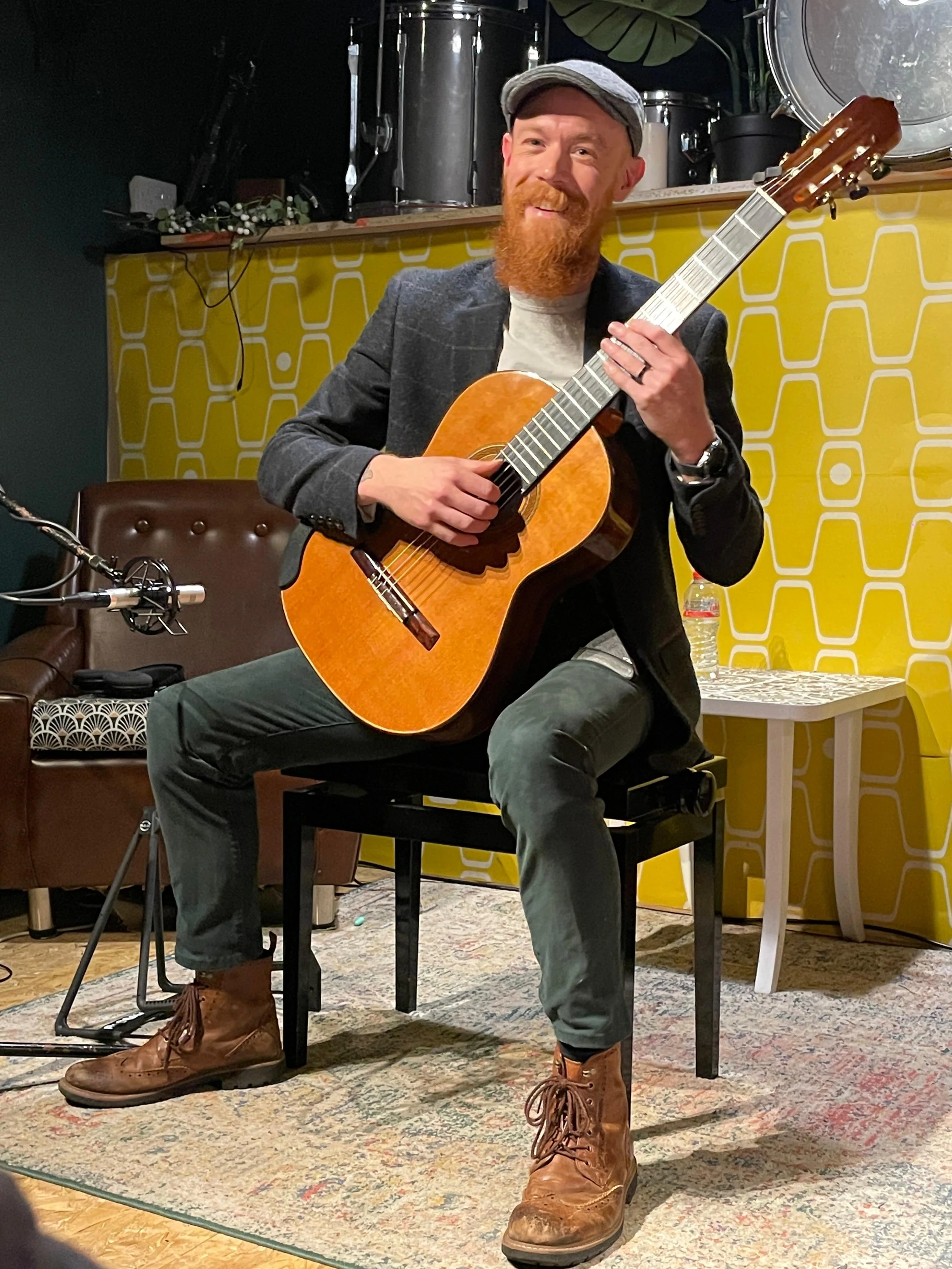 A man with a red beard, gray cap, gray blazer, light T-shirt, dark pants, and brown boots is sitting on a chair playing an acoustic guitar indoors. There is a microphone and sound equipment nearby, and the background has a yellow patterned wall and some musical instruments.