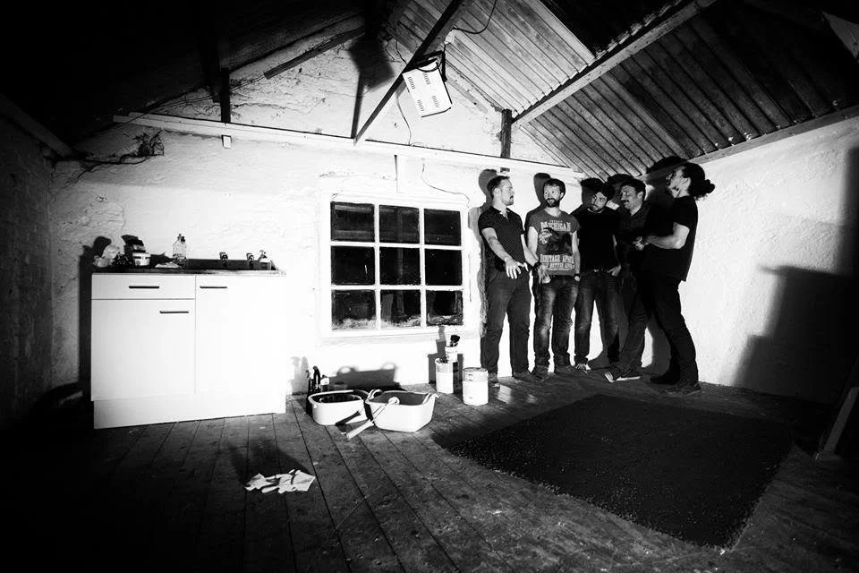 Five people standing inside a rustic, unfinished attic room with a sloped ceiling. They are gathered near a window, and various painting supplies are on the floor and against the wall. The room has a wooden floor and a small cabinet on the left.