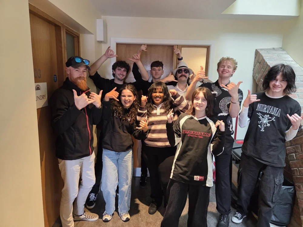 Group of nine young people indoors, making hand gestures, smiling, wearing casual and band-themed clothing, with a brick fireplace and a wooden door in the background.