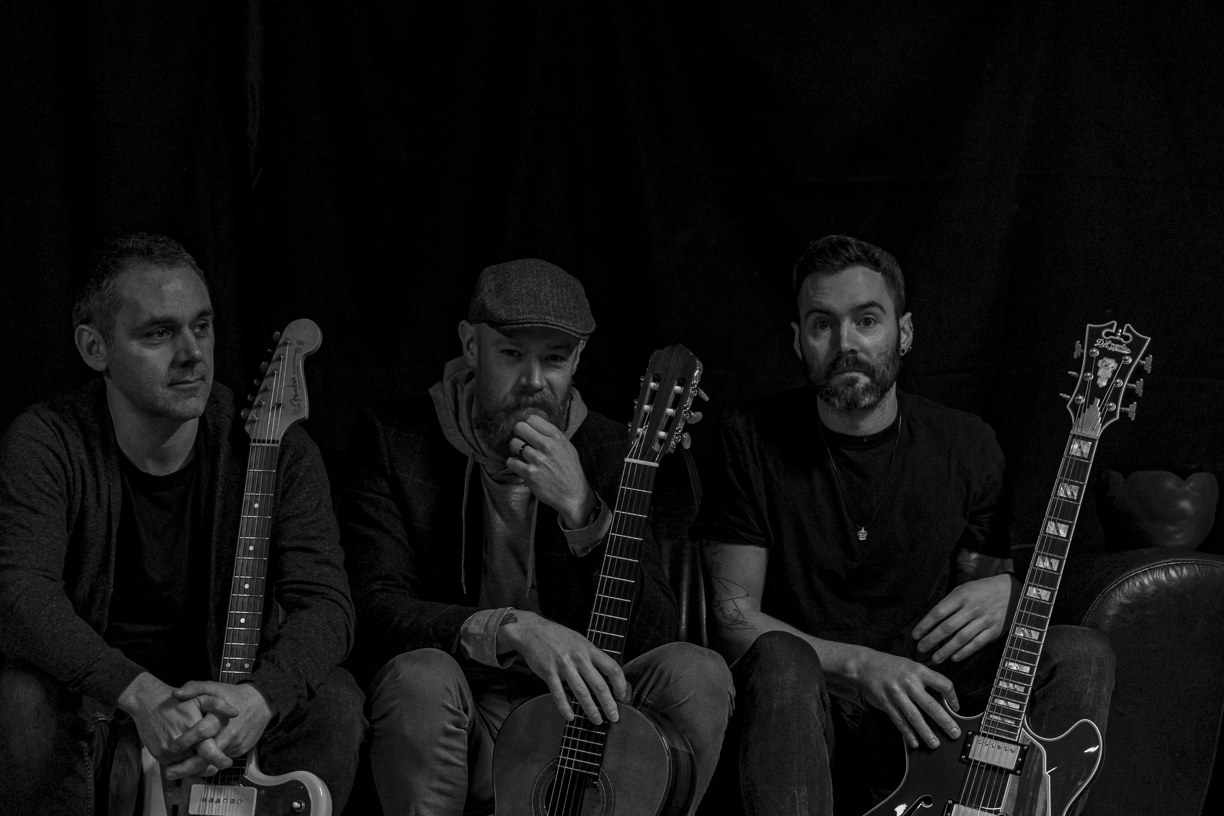 Four men sitting in a row, each holding or positioned with a guitar, against a dark background. The men vary in appearance, with one wearing a flat cap, others have different hairstyles and facial hair, and expressions range from serious to contemplative.