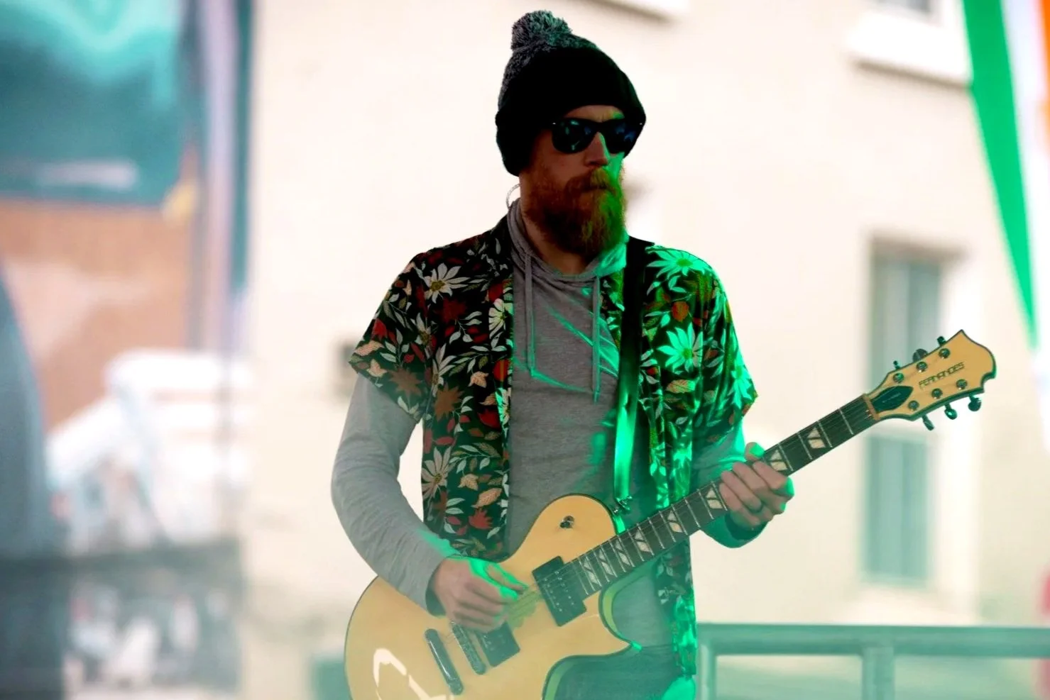 A man with a beard wearing sunglasses, a black beanie, a gray hoodie, and a floral shirt playing a cream-colored electric guitar. Green stage lighting is cast on him.