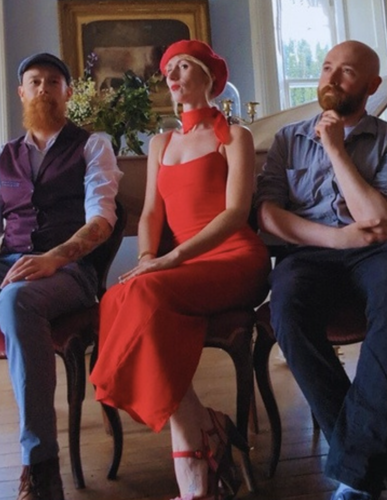 Three people sitting on chairs in a room with a large window and a framed painting behind them. The woman in the center is wearing a red dress, matching red hat, and appears to be posing confidently. The men on either side have relaxed postures, one with a beard and the other with a mustache and beard.