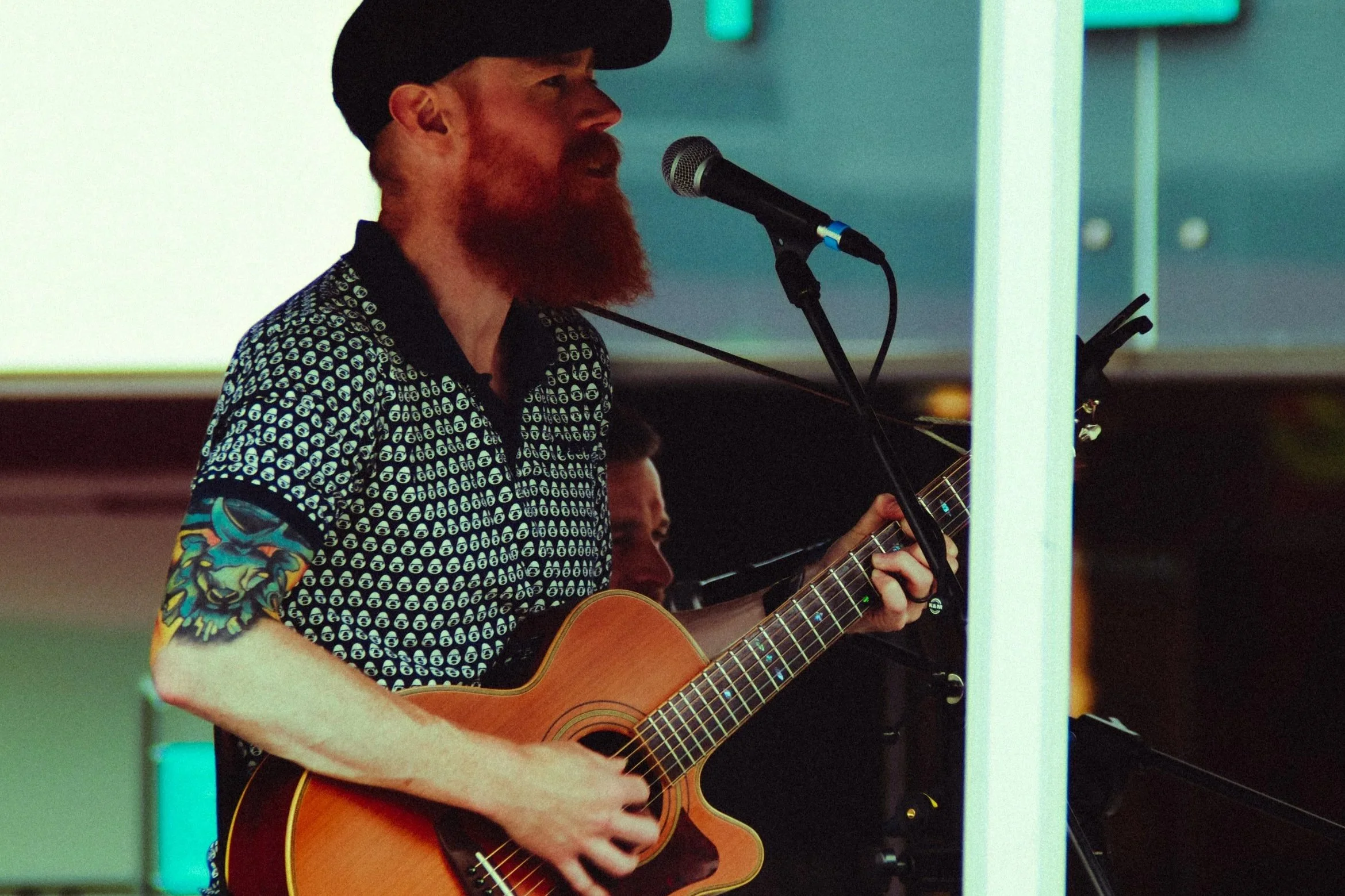 A man with a red beard, tattoos on his arm, wearing a black cap and a patterned shirt, playing an acoustic guitar and singing into a microphone.