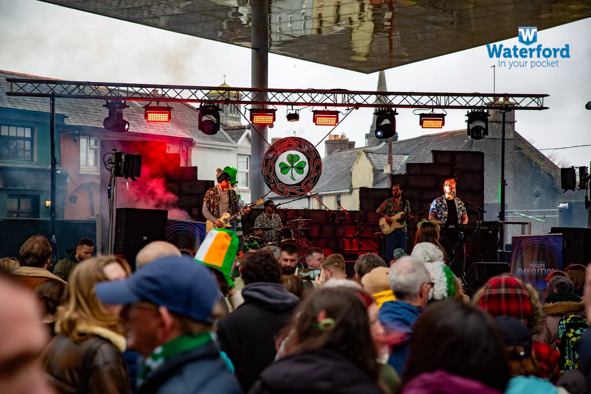A music band performing live on an outdoor stage during a festival with a crowd gathered in front. The scene includes stage lights, musical instruments, and a backdrop featuring the Irish shamrock. People in the audience are wearing festive attire, including a notable rainbow-colored hat.