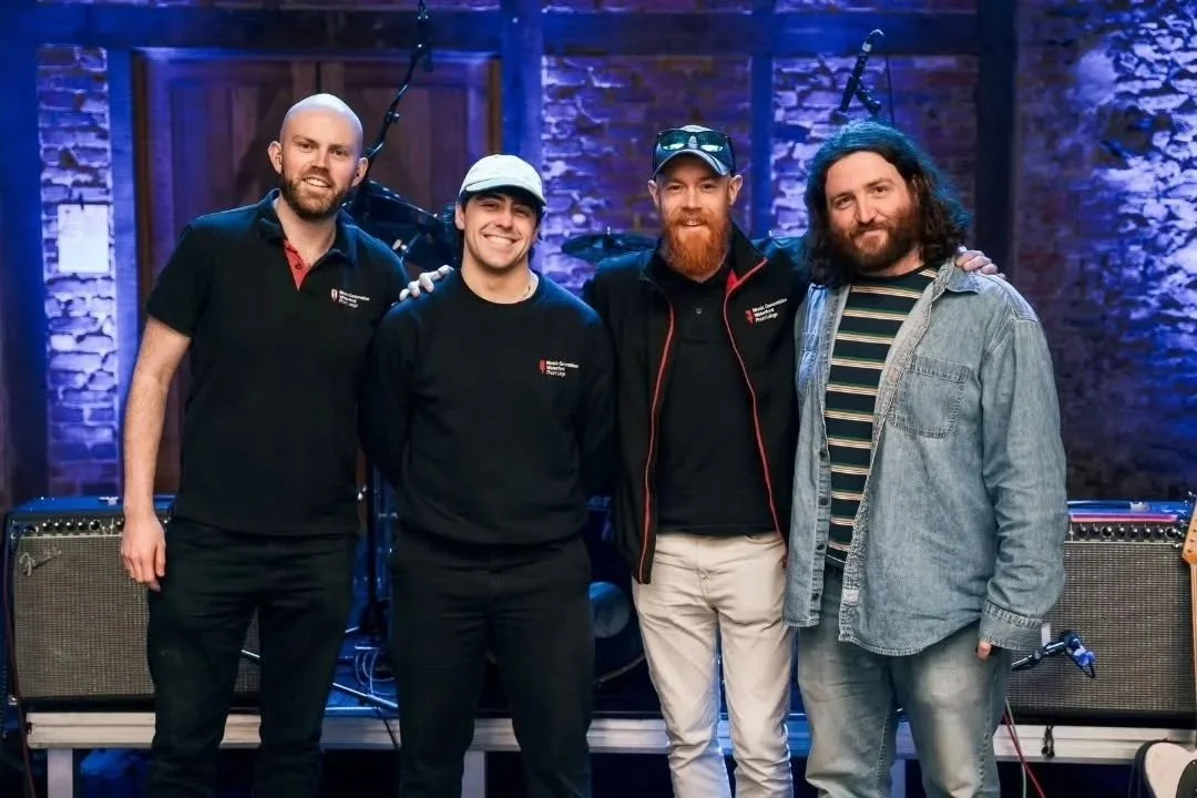 Four men standing together in a music studio or stage setting, smiling at the camera. Two amplifiers are visible in the background, with exposed brick walls and stage lighting.