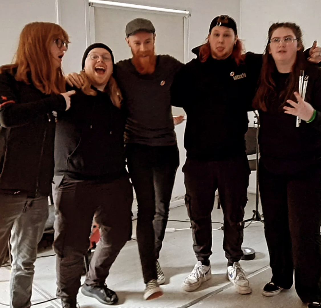 Five people standing together indoors, smiling and posing for a group photo, with arms around each other's shoulders.