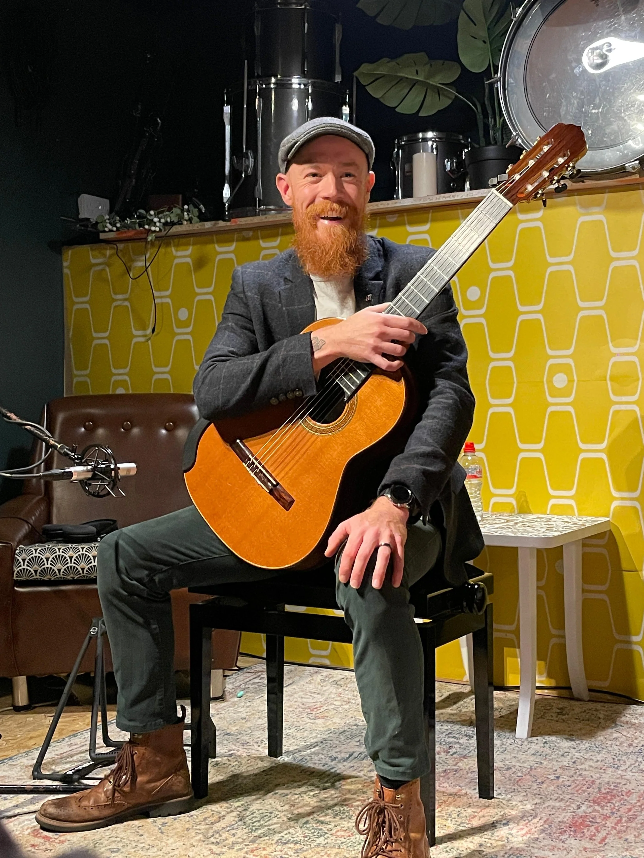 A man with a red beard, wearing a gray flat cap, dark blazer, and leather boots, is sitting on a chair holding an acoustic guitar. He is smiling and looking at the camera, with a drum set and green plant leaves in the background.