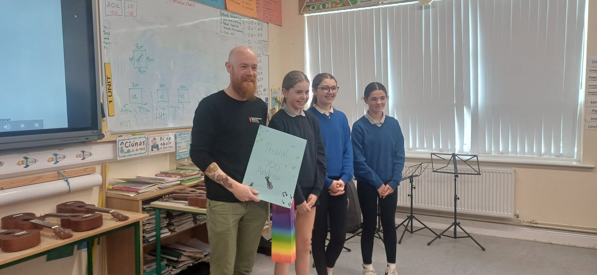 A man wearing a Black T-shirt with a tattoo on his arm is holding a colorful sign that says 'Thank you Andrew' and standing with three young girls dressed in school uniforms next to a whiteboard in a classroom.