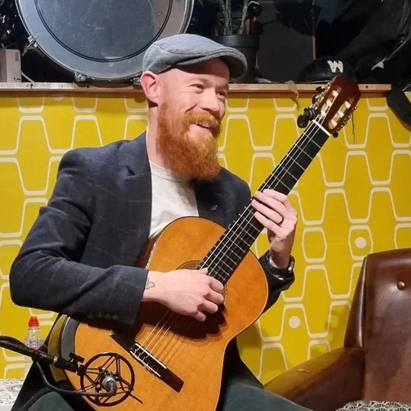 A man with a red beard and mustache, wearing a gray flat cap and a dark blazer, plays an acoustic guitar and smiles. He is sitting in a room with yellow patterned wallpaper.