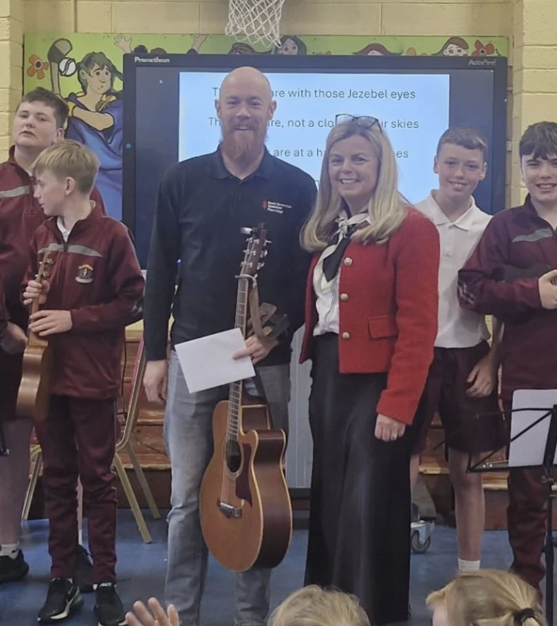 Group photo of children and two adults, one man holding a guitar, in a school gymnasium with a colorful mural and a basketball hoop in the background.
