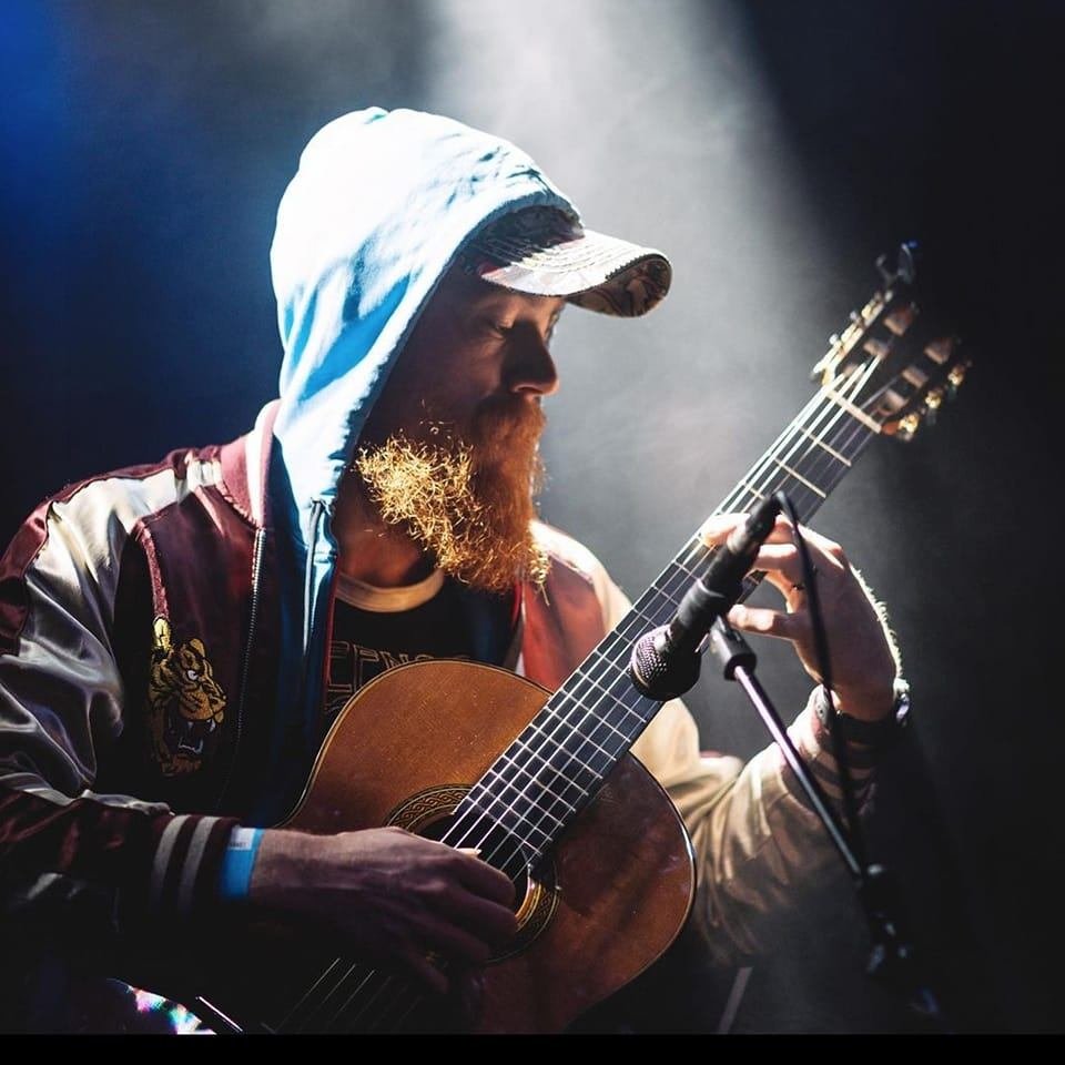 Man with a beard wearing a white hoodie and a cap playing an acoustic guitar on stage.