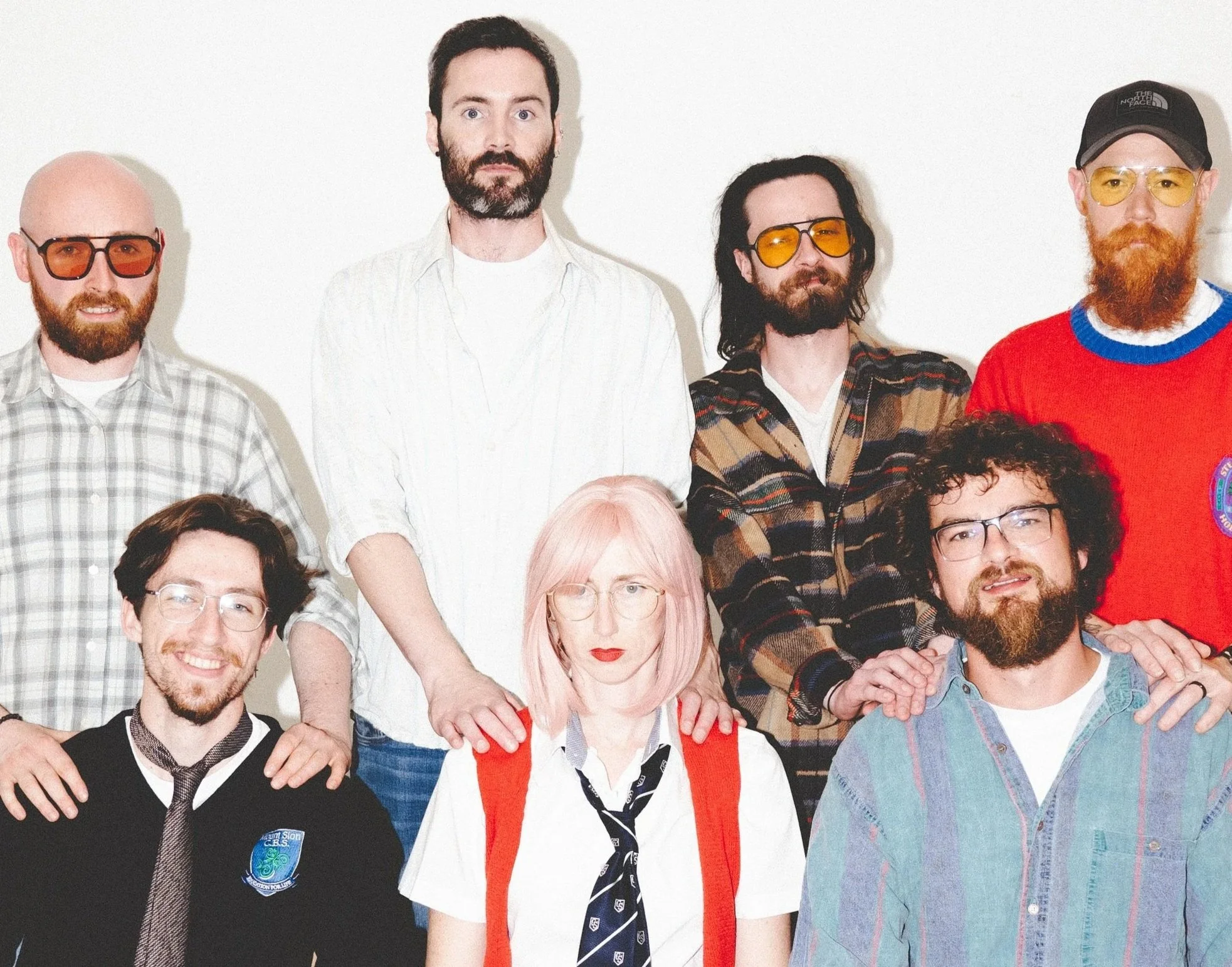 A group of diverse people posing for a photo against a plain white background, some wearing glasses and casual attire, with one woman with pink hair in front.