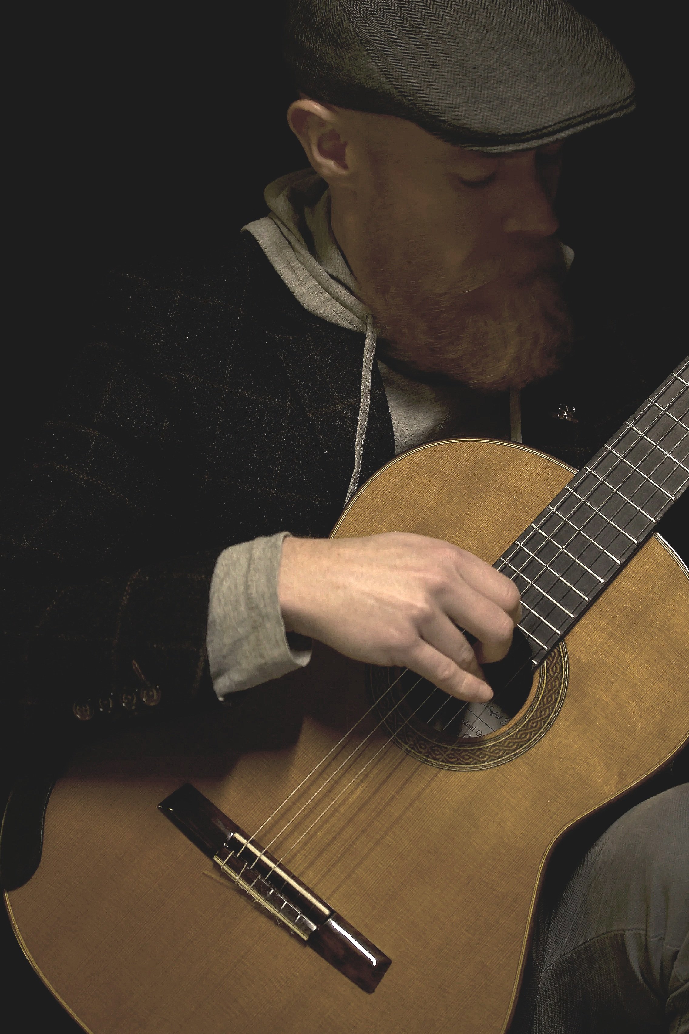A man with a beard wearing a gray flat cap and a hoodie, playing an acoustic guitar.