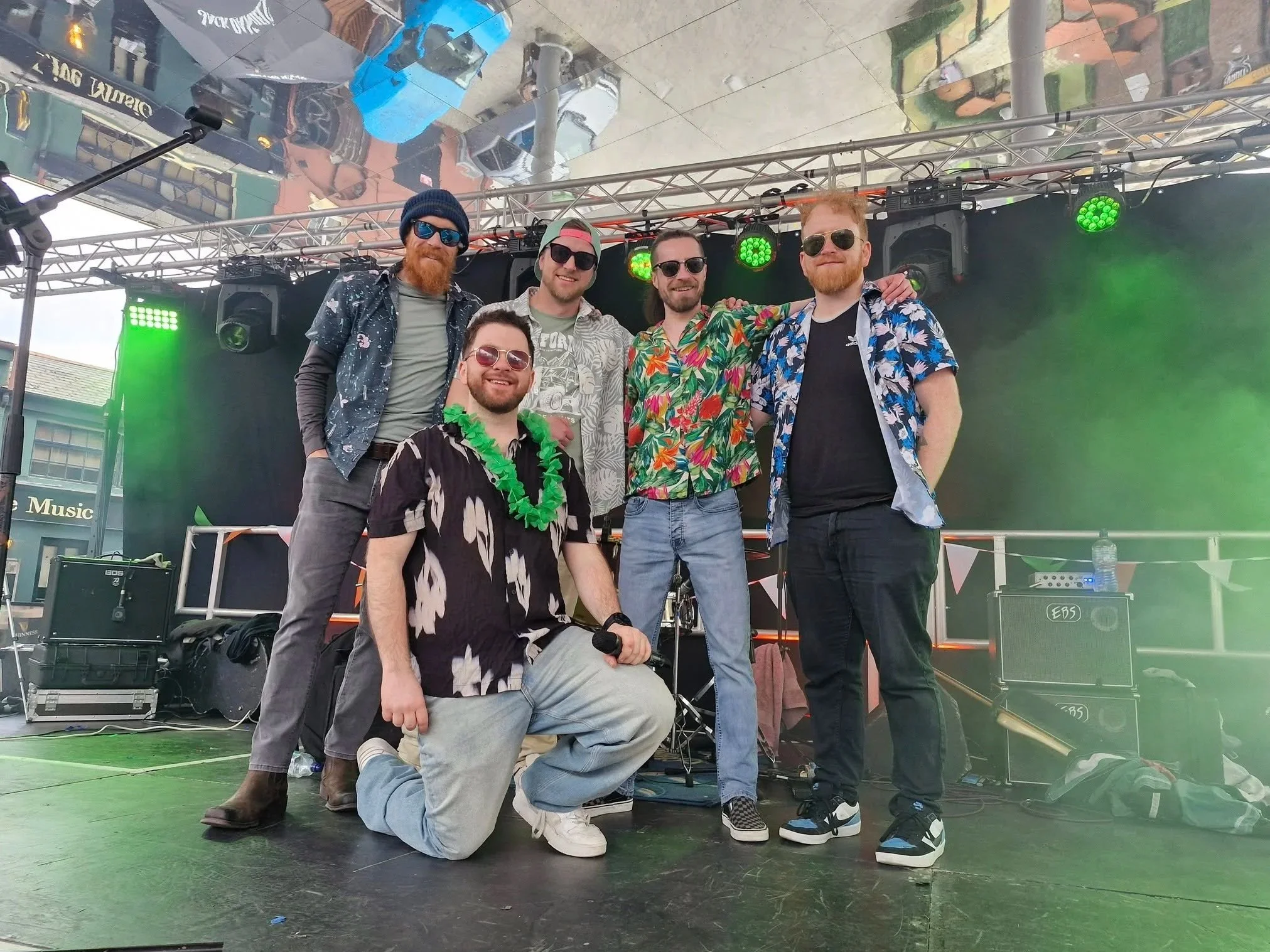 Group of six men on a stage outdoors, smiling, wearing casual and tropical-themed clothing, some with sunglasses and hats, with musical equipment and green stage lights in the background.