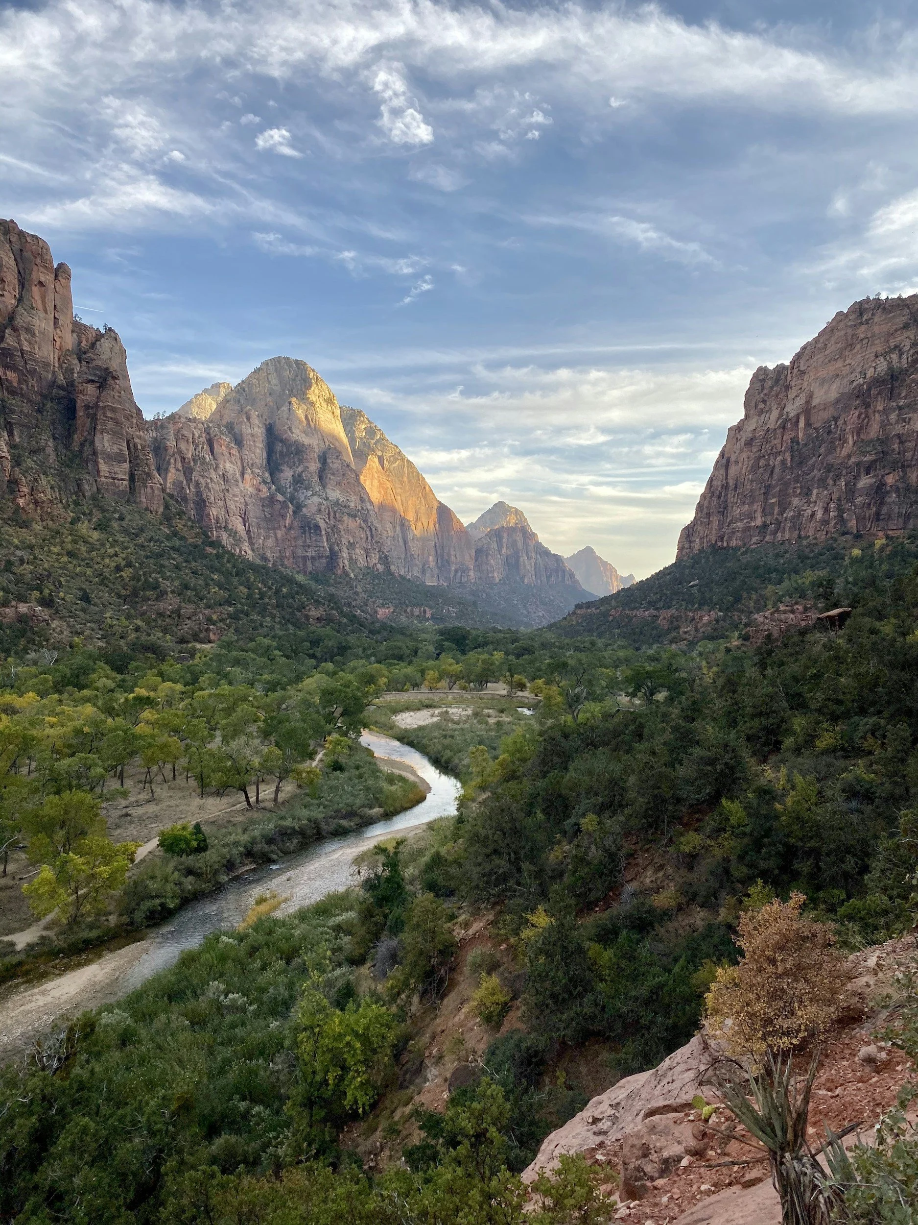 Scenic view of a river flowing through a valley surrounded by tall rocky mountains and lush green trees, under a partly cloudy sky.
