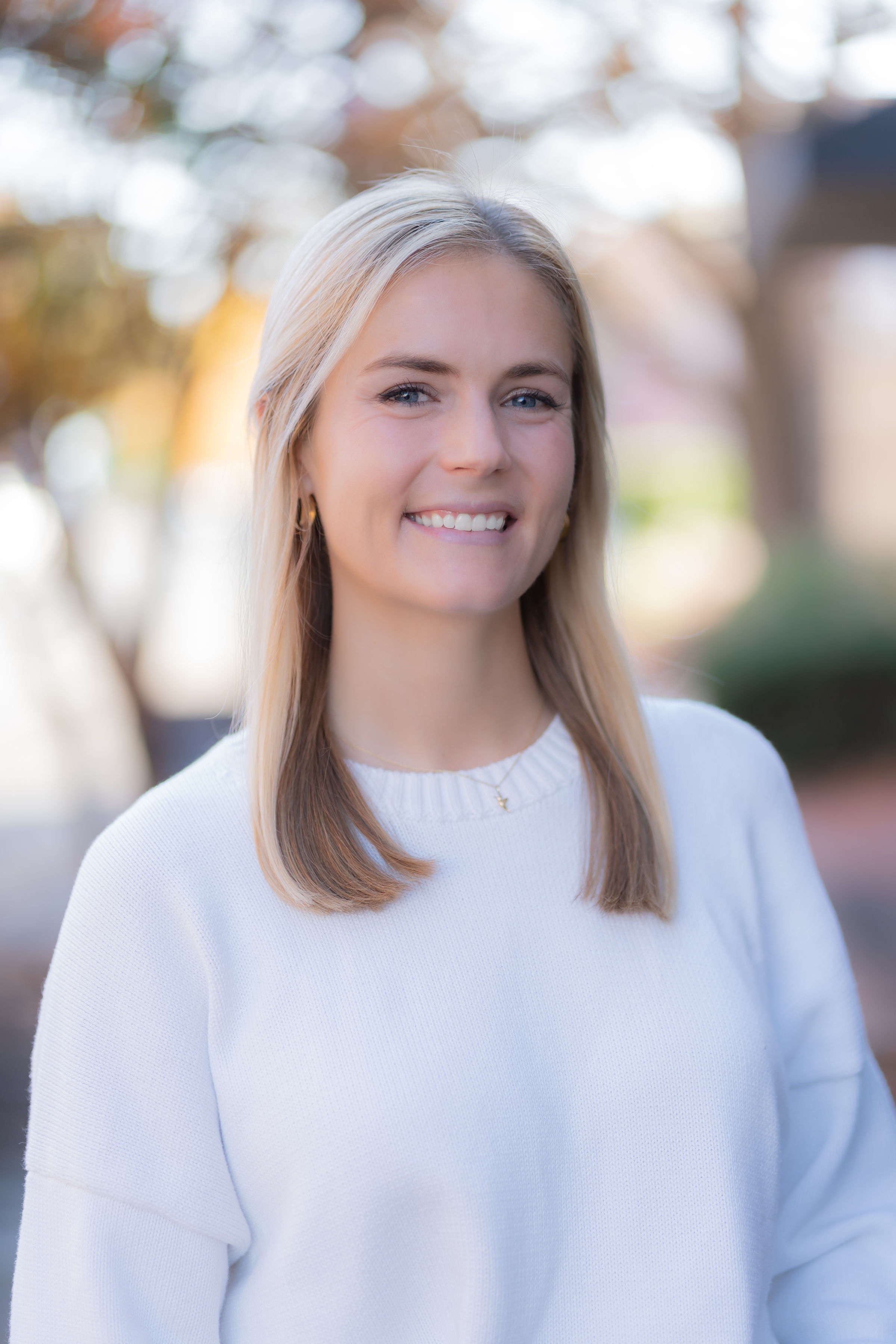 A young woman with blonde hair and blue eyes smiling outdoors in a white sweater.