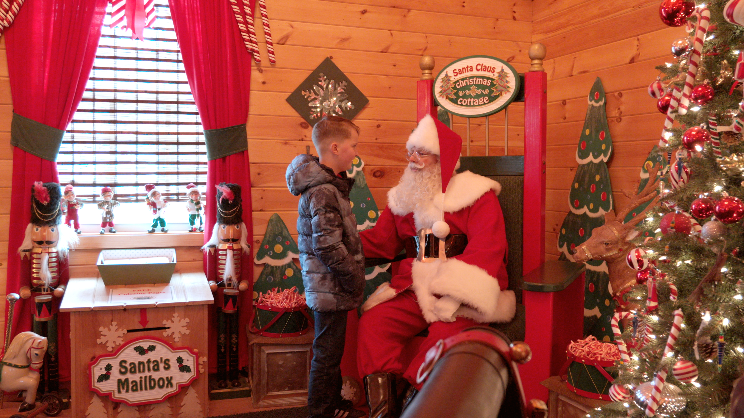 A young boy talking to Santa Claus, who is dressed in a traditional red suit with white fur trim, inside a decorated Christmas booth at Santa's Mailbox. The booth has wooden walls decorated with Christmas trees, nutcracker figures, and holiday orname