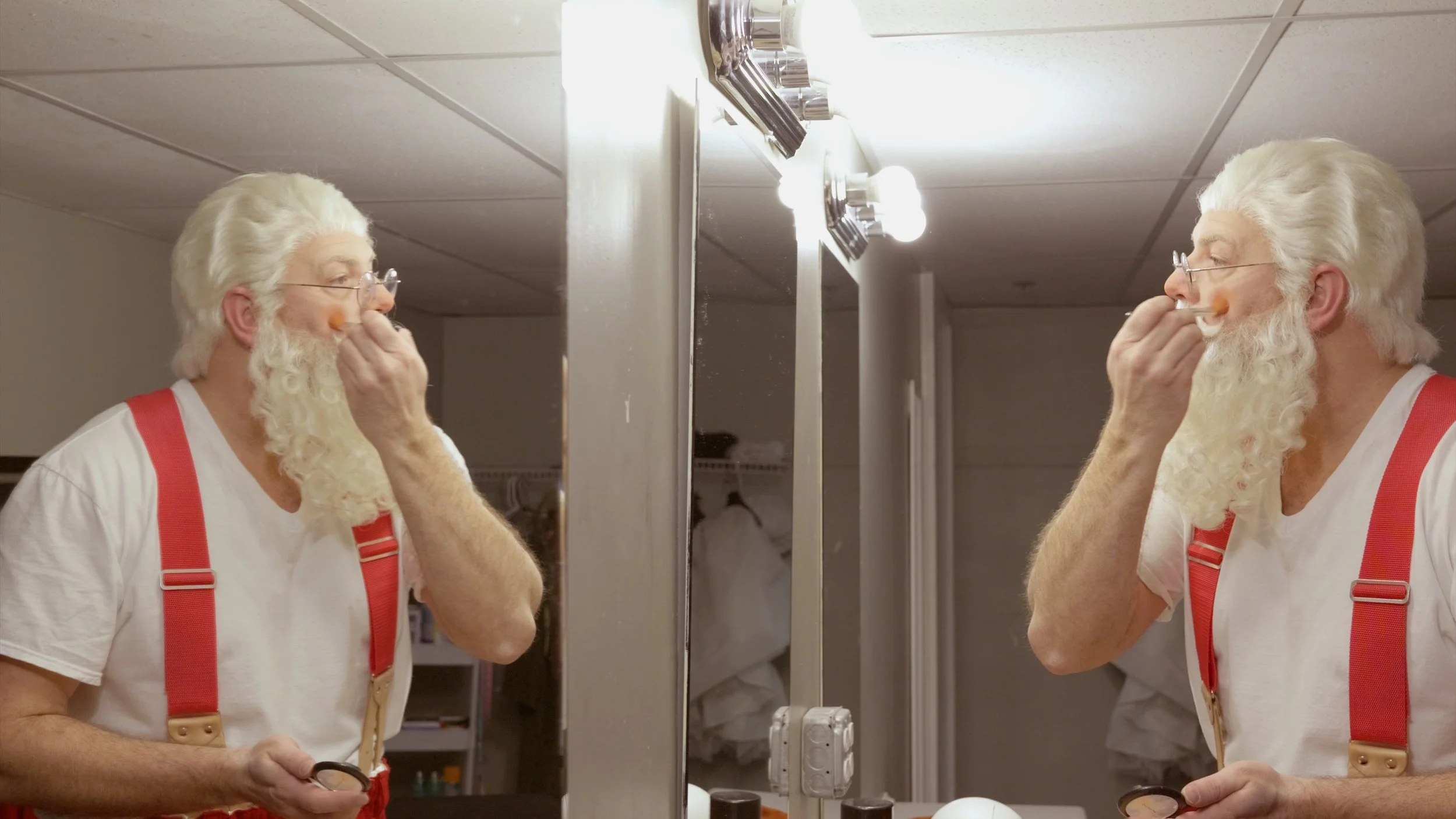 An elderly man with white hair, glasses, and a beard applies makeup in front of a mirror in a dressing room. He is wearing a white shirt with red suspenders. The reflection shows him carefully applying makeup with a brush.