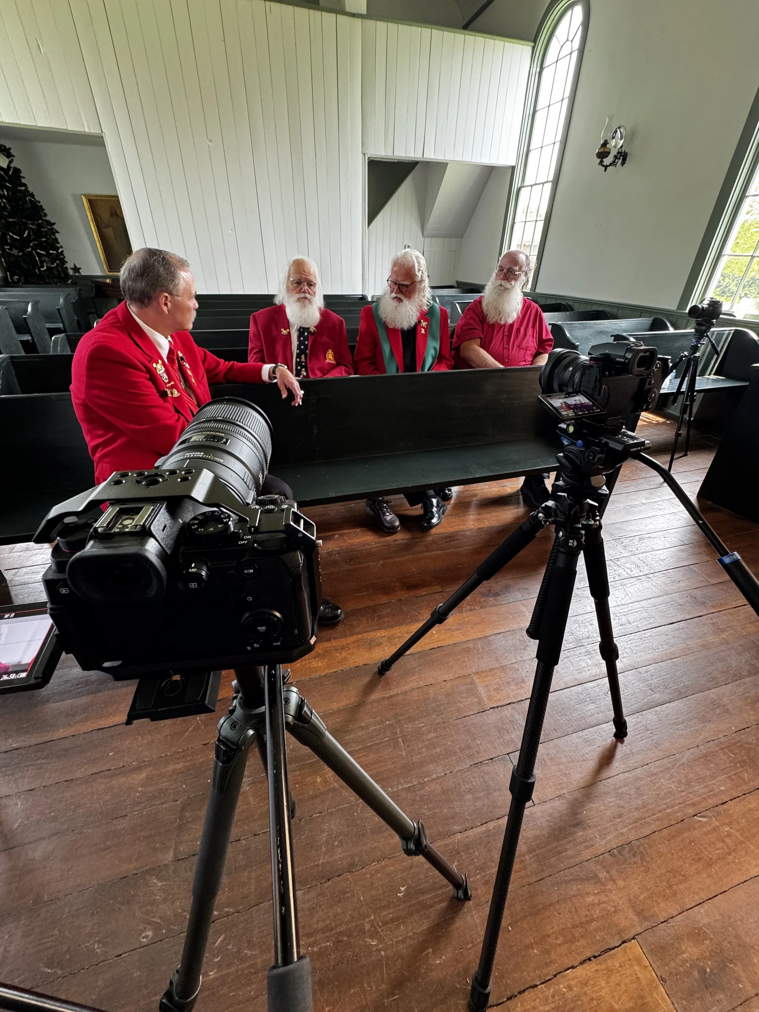 Four men dressed in red jackets, some with white beards, sitting in a church pew inside a church, with camera equipment set up in front of them.