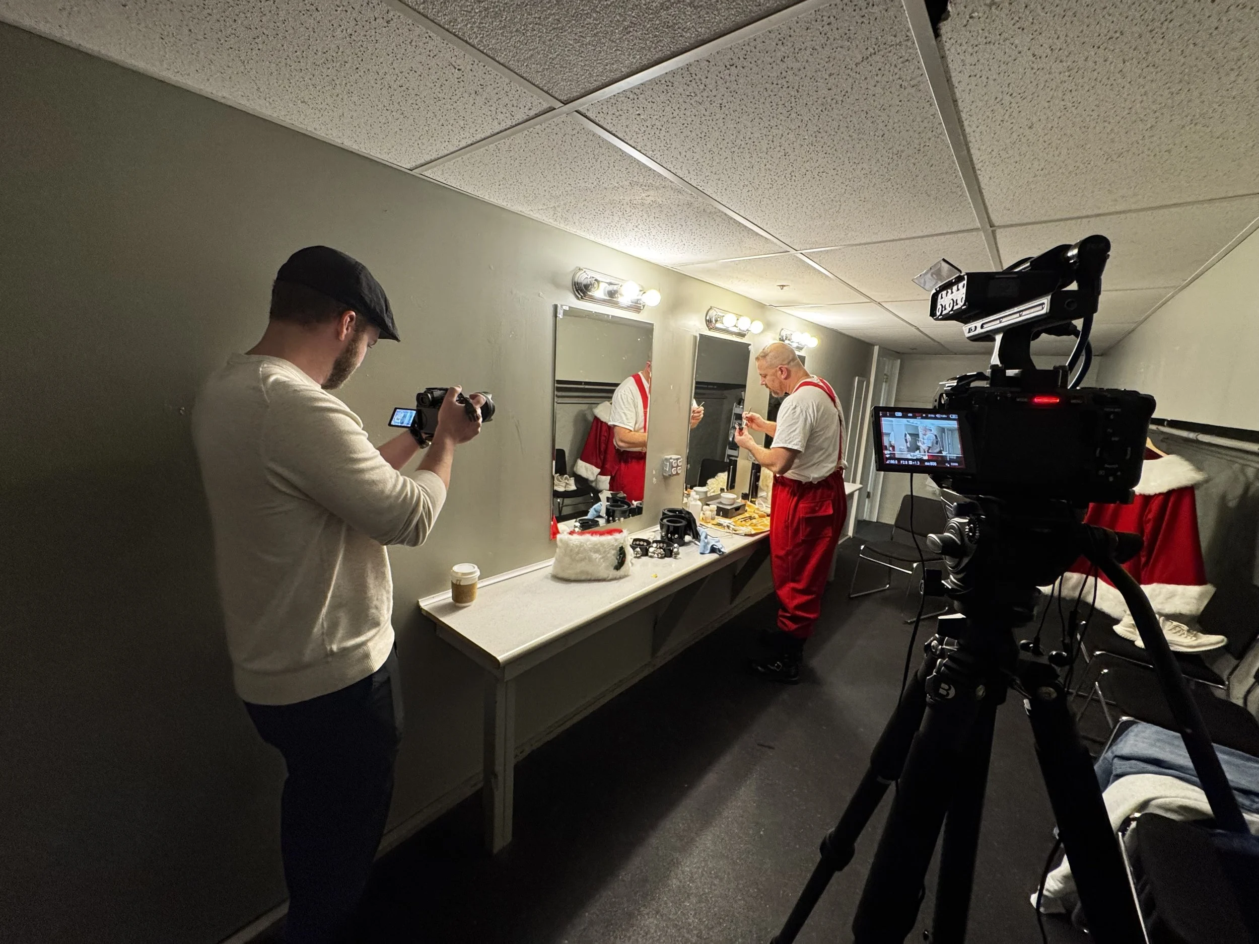 A behind-the-scenes view of a filming crew capturing footage of a man preparing for a Christmas-themed scene, as indicated by Santa hats and holiday decorations on a counter and the presence of a professional video camera.