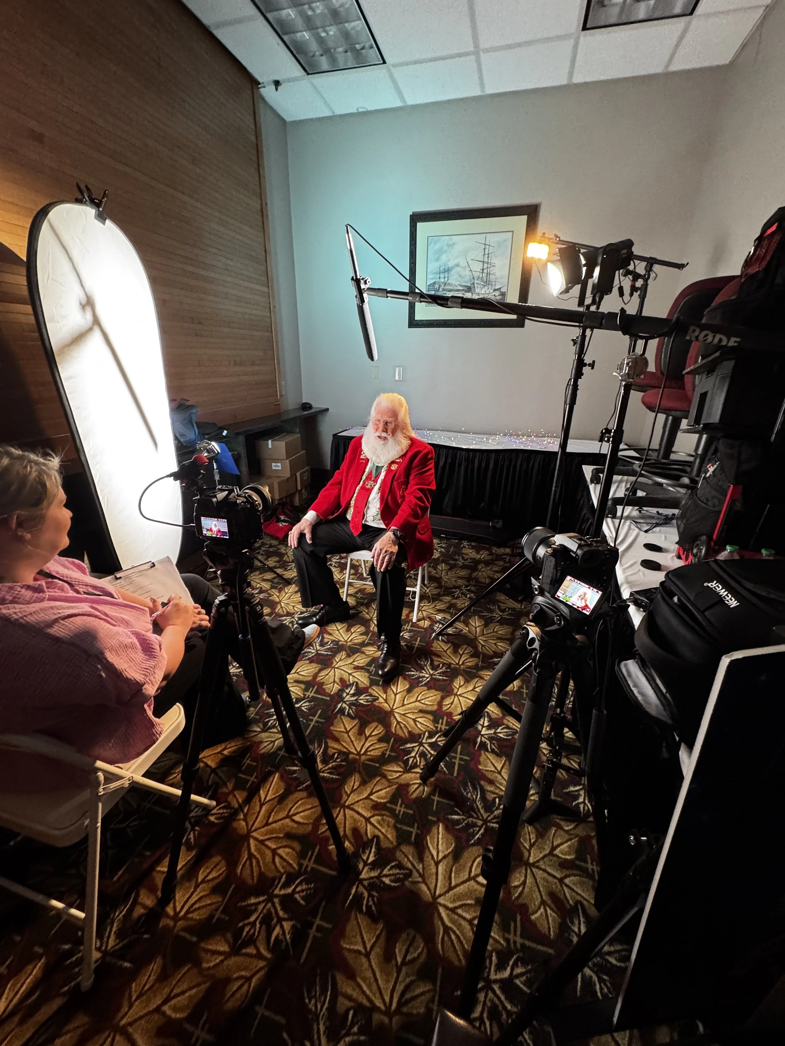 A man dressed as Santa Claus sitting on a chair in a room being filmed or photographed. Lights, cameras, and equipment are set up around him, with a woman seated nearby holding a script or notes. The room has a patterned carpet, a framed picture of ships on the wall, and various filming gear is visible.