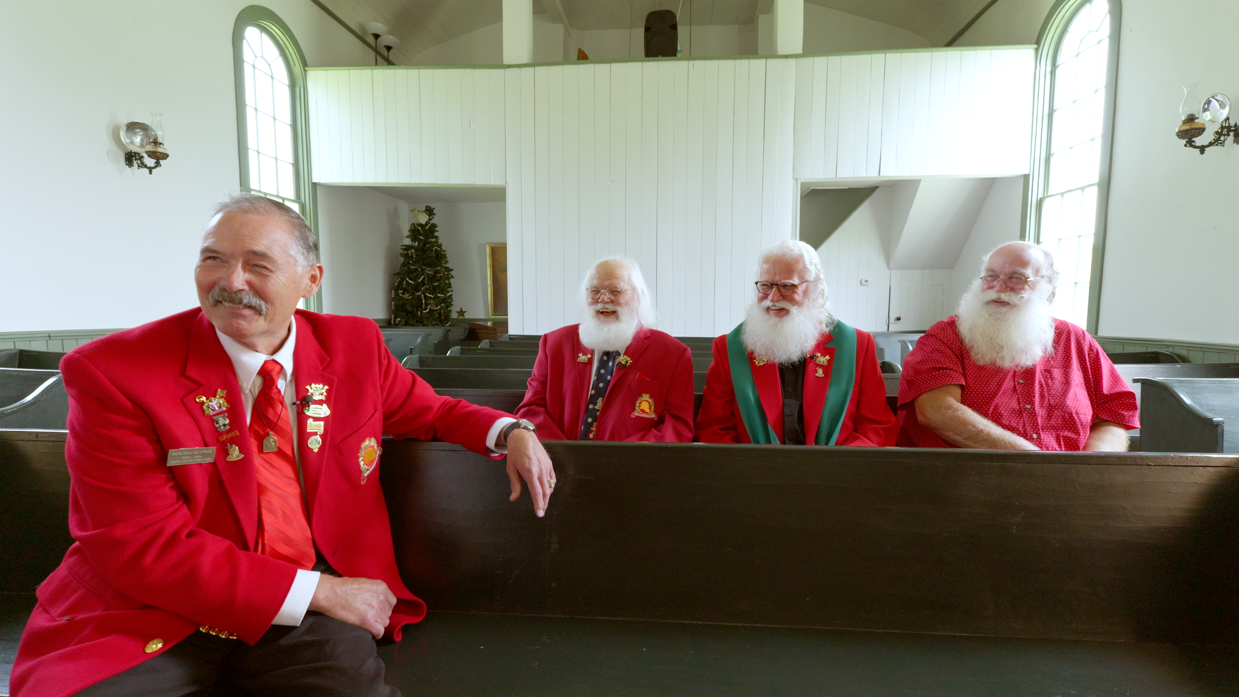 Four elderly men with white beards and glasses sitting in church pews, dressed in red and green holiday attire, inside a church with large windows and white walls.