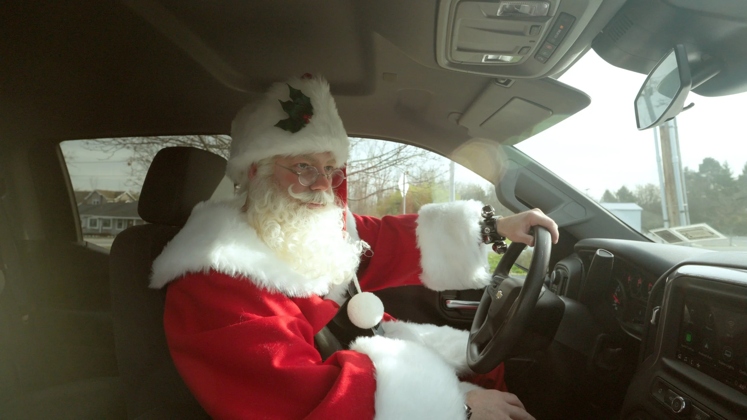 Man dressed as Santa Claus driving a vehicle, wearing a Santa suit, hat with holly decoration, and glasses.