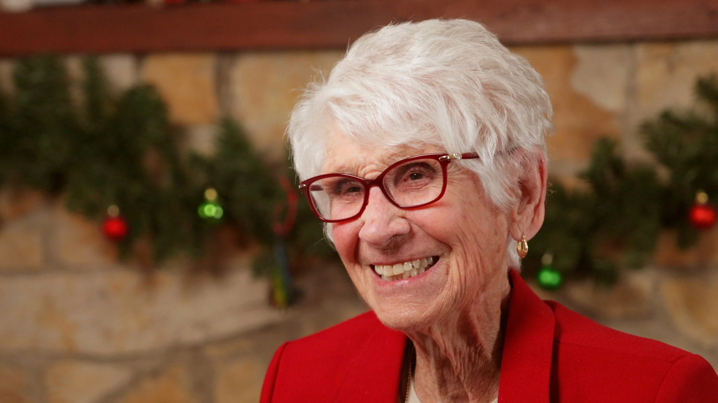 Close-up of elderly woman with white hair wearing red glasses and a red jacket, smiling, with a decorated Christmas tree in the background.