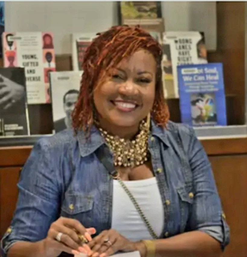 A woman with reddish braids, smiling, wearing a denim shirt and a layered pearl necklace, sitting at a table with books in the background.