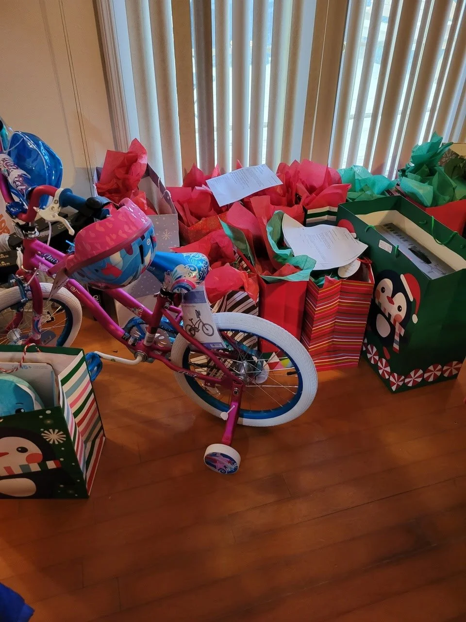 A pink children's bicycle with training wheels and a snowflake-patterned helmet, surrounded by Christmas gift bags and presents on a hardwood floor near a window with vertical blinds.