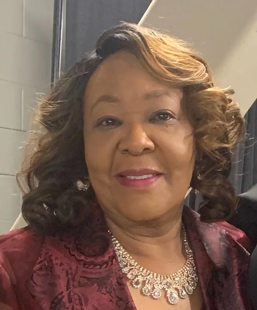 A woman with shoulder-length, curly hair smiling at the camera, wearing a maroon patterned jacket and a sparkling necklace.