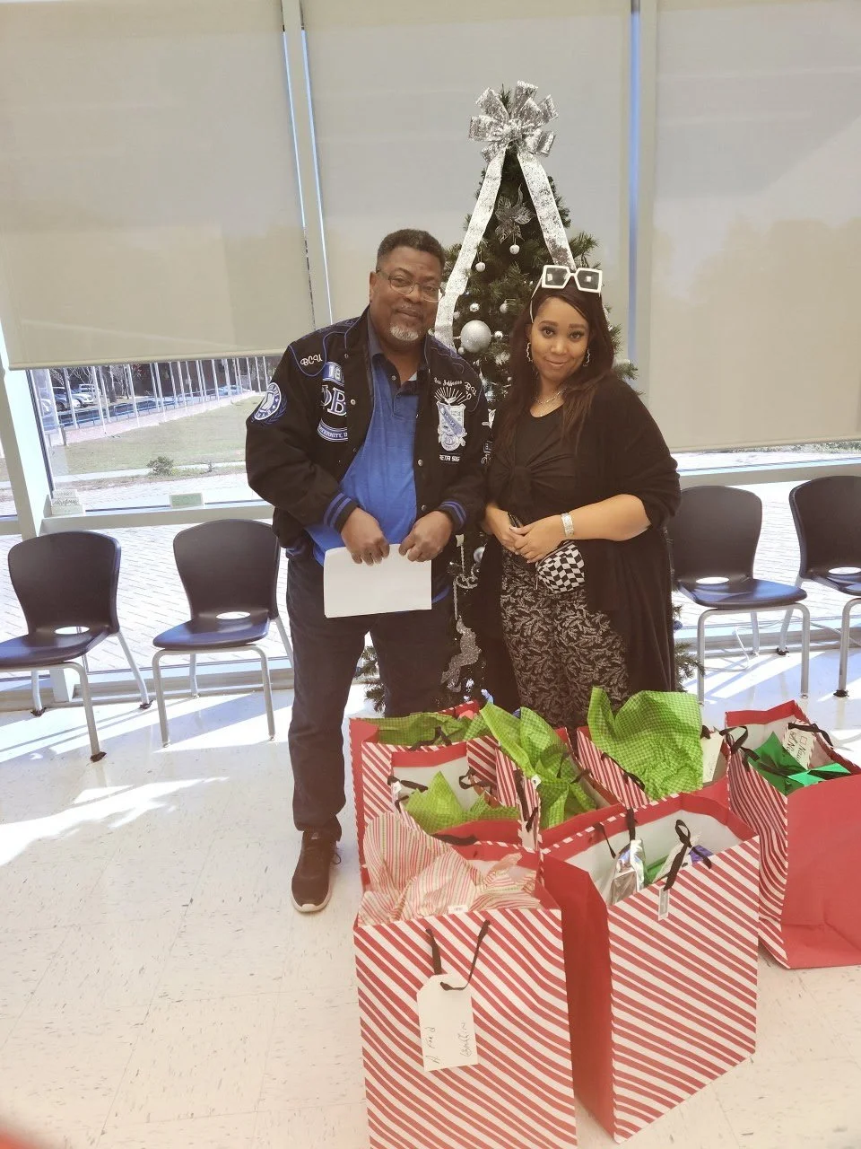 Two people standing in front of a decorated Christmas tree, with gift bags on the floor in front of them, in a room with large windows.
