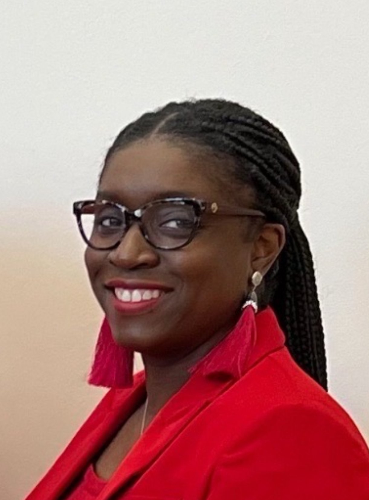 A woman smiling, wearing glasses, earrings, and a red blazer with braids in her hair, standing against a plain white background.