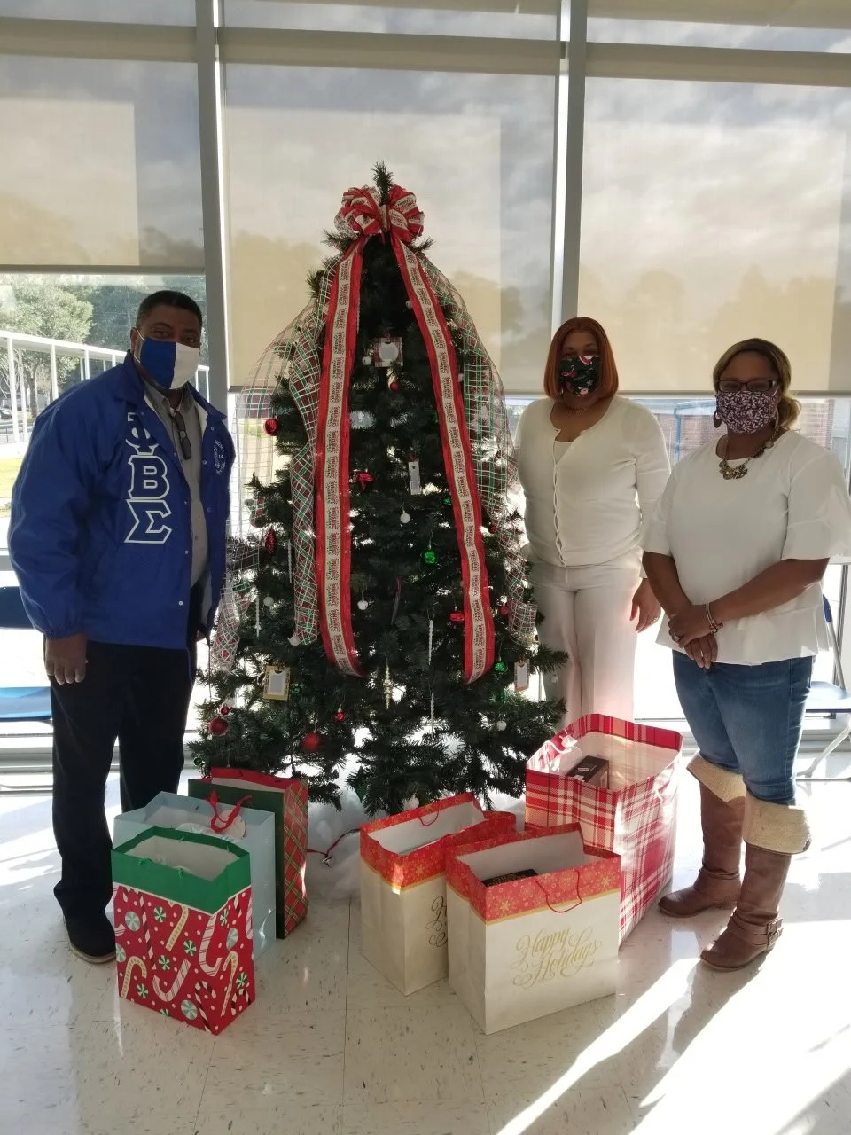 Three people standing next to a decorated Christmas tree with wrapped presents underneath in a bright indoor space.
