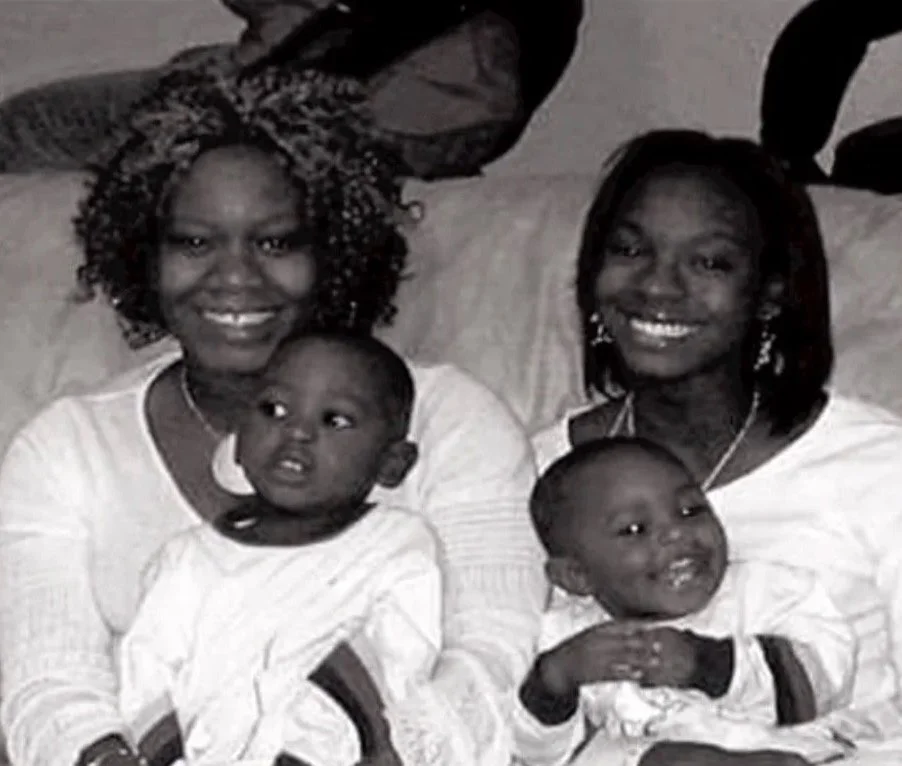 Two women with curly and straight hair smiling, each holding a young child, on a couch with decorative balloons in the background.