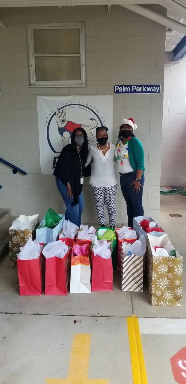 Three women wearing masks standing in front of a school banner and a sign that says Palm Parkway, surrounded by wrapped Christmas gift bags.