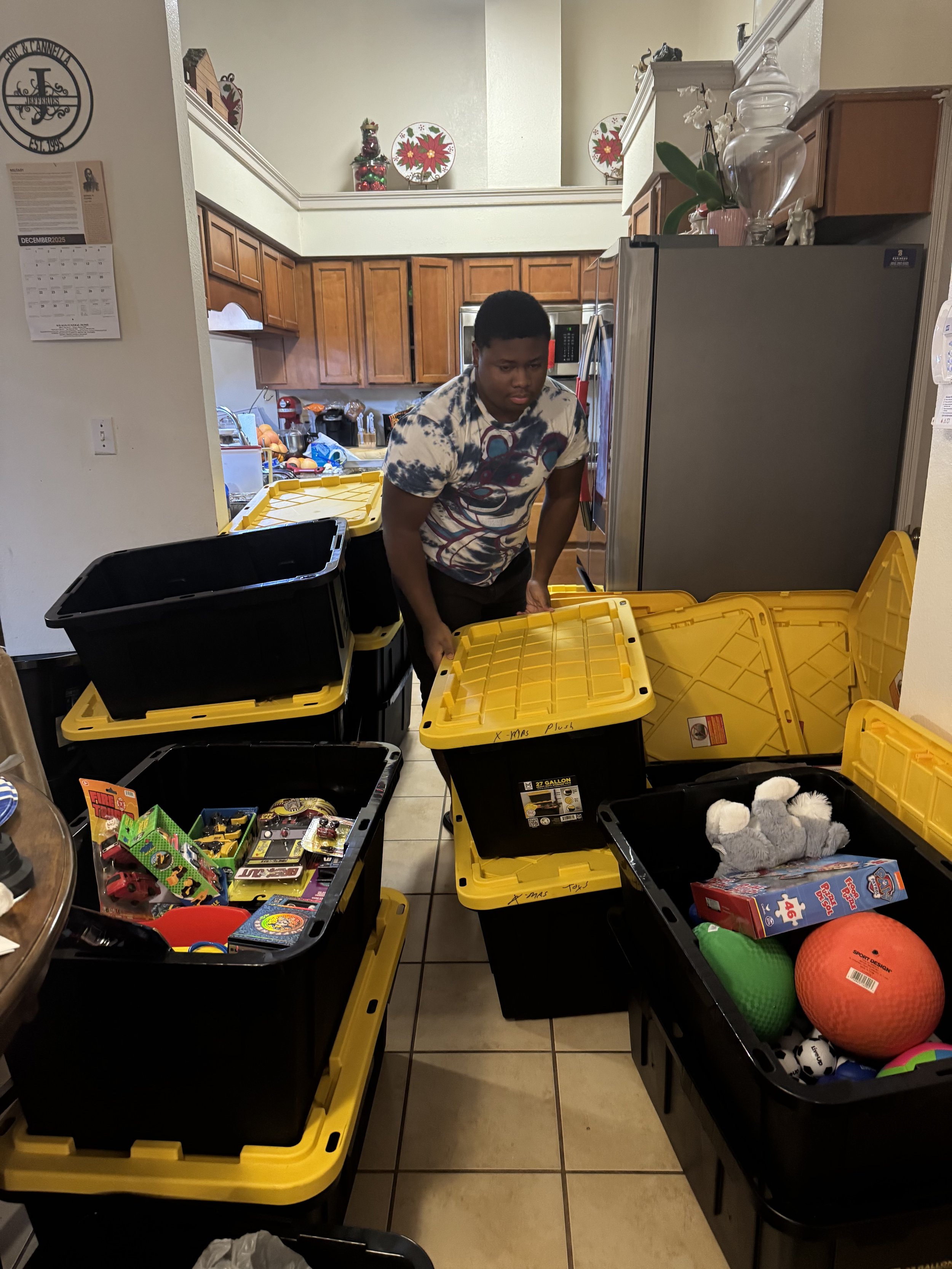 A young man packing or sorting toys in plastic bins in a kitchen. The bins contain various toys and sports equipment, with Christmas decorations visible on the upper shelf.